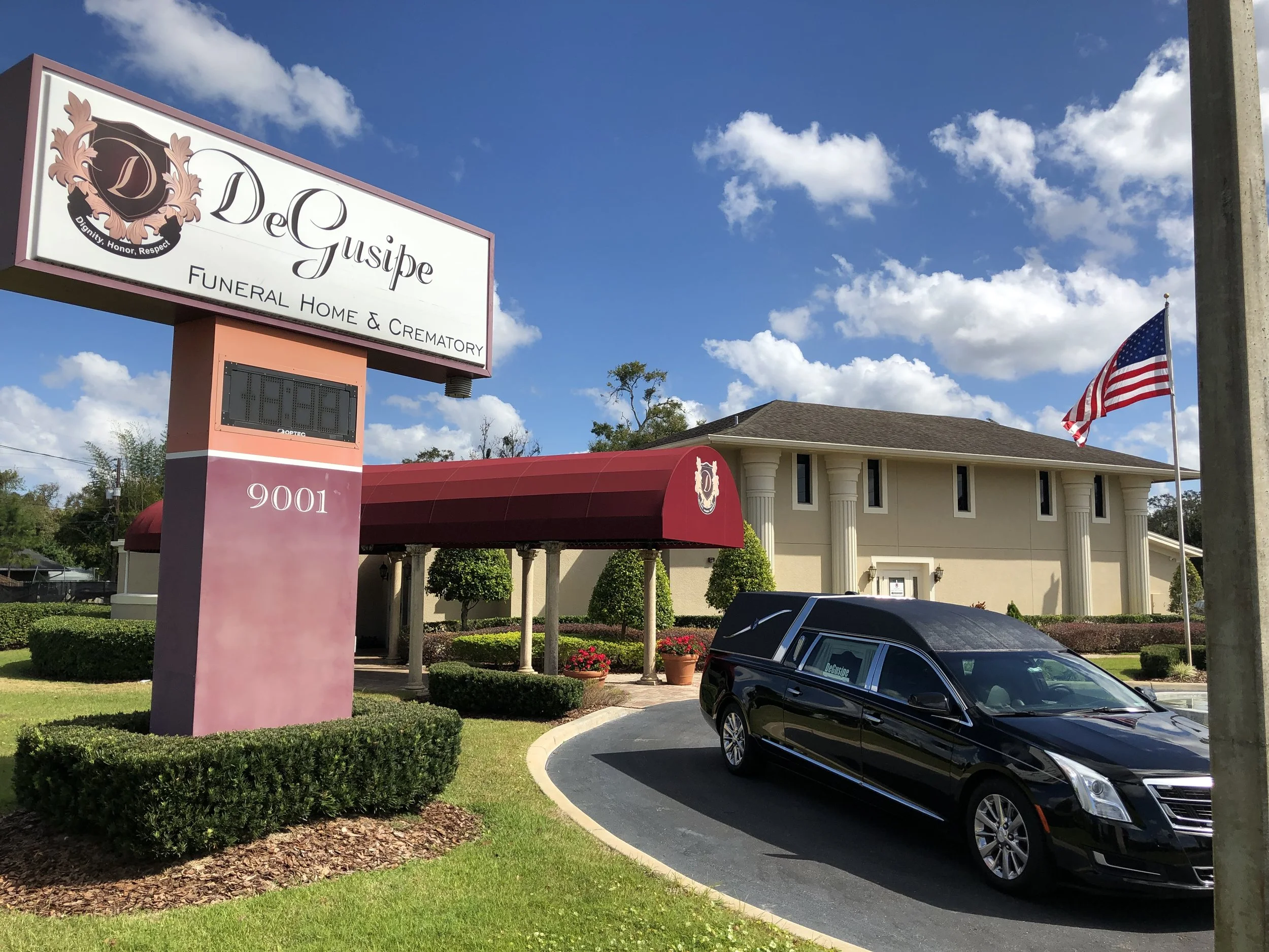 DeGuisse Funeral Home & Crematory building with a large sign, a black hearse in the parking lot, a flagpole with an American flag, and a blue sky with clouds.
