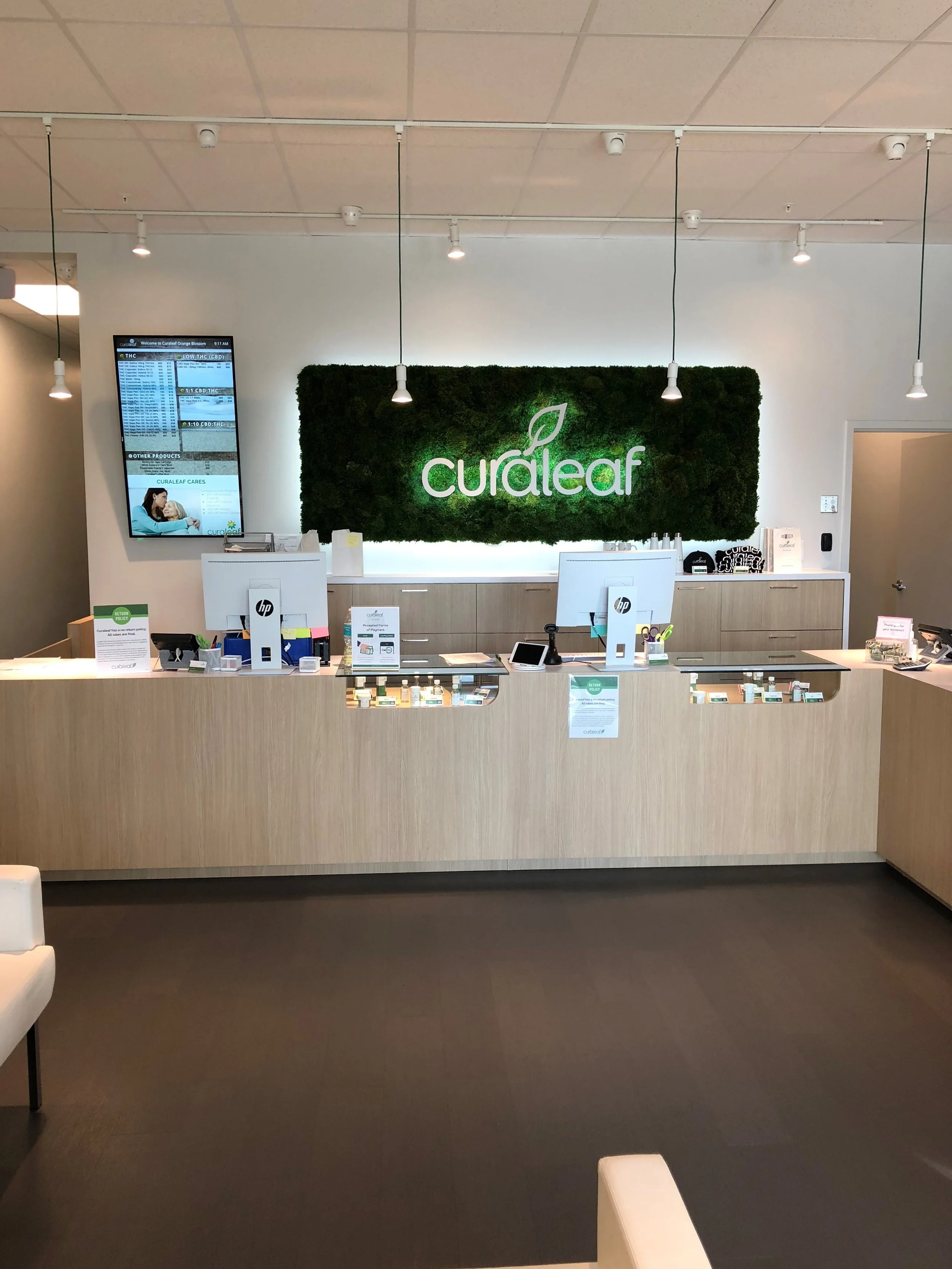 Interior of a reception area with a large illuminated sign reading 'cureleaf' against a green moss wall, behind a light wood counter with computers and display cases, and a digital screen on the left showing various information.