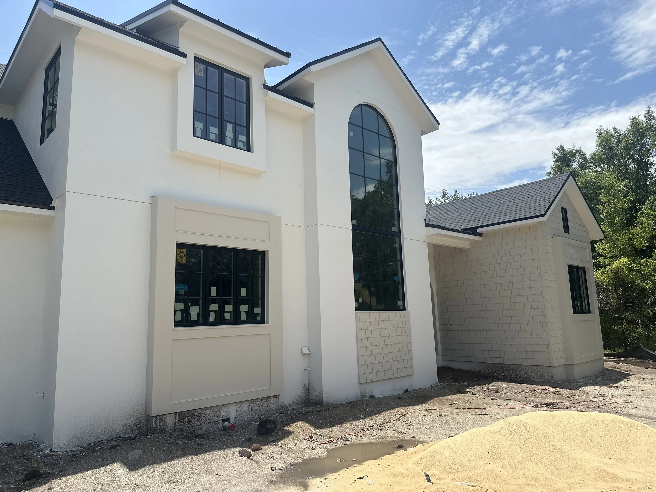New two-story house under construction with white exterior walls, black window frames, and a large arched window, surrounded by dirt and construction materials, with a partly cloudy sky in the background.