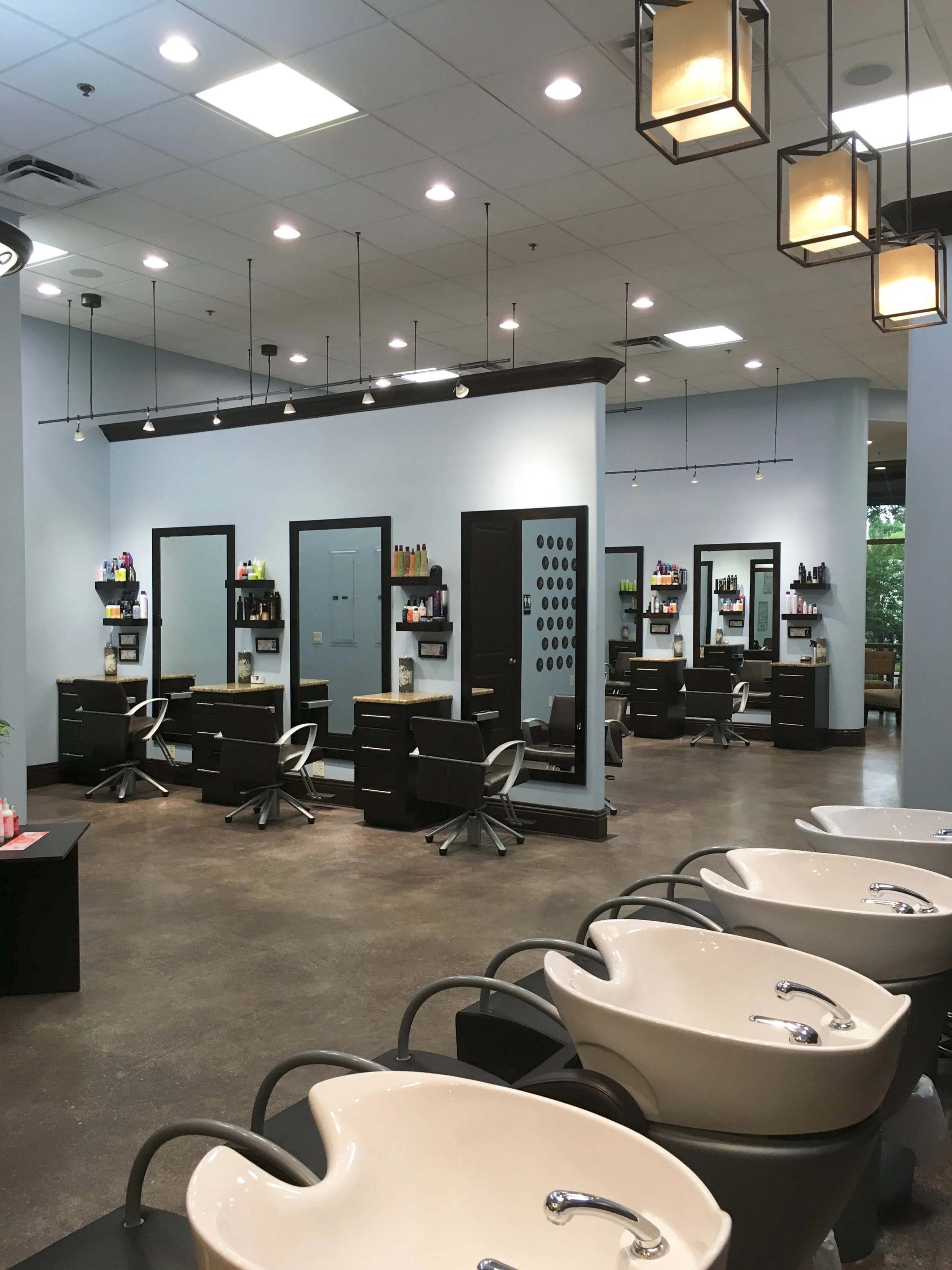 Hair salon interior with styling stations, mirrors, chairs, and shampoo bowls.