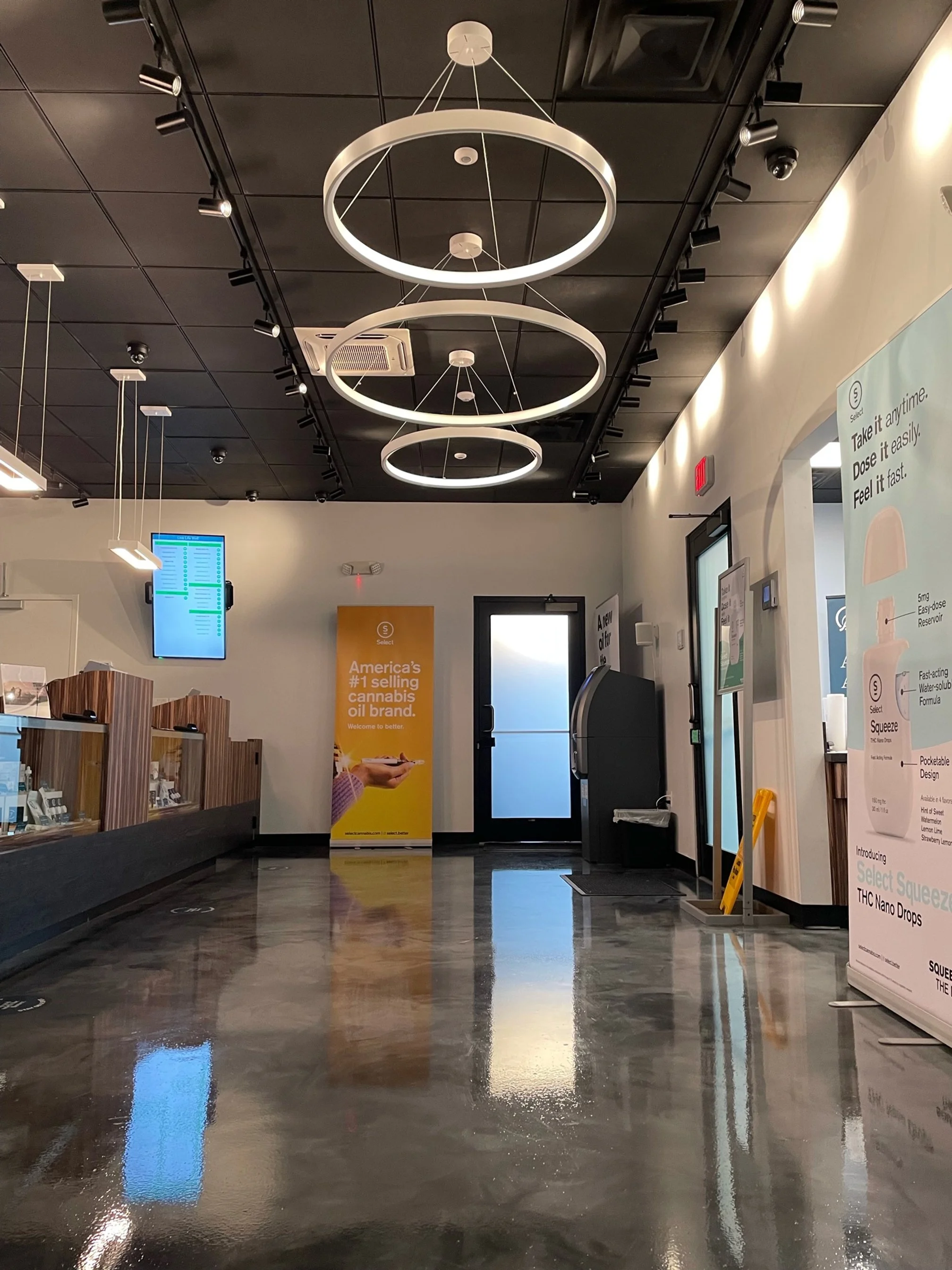 Interior of a modern cannabis dispensary with polished concrete floors, black ceiling with ring-shaped light fixtures, a digital menu display, and promotional banners for THC nano drops and cannabis oil.