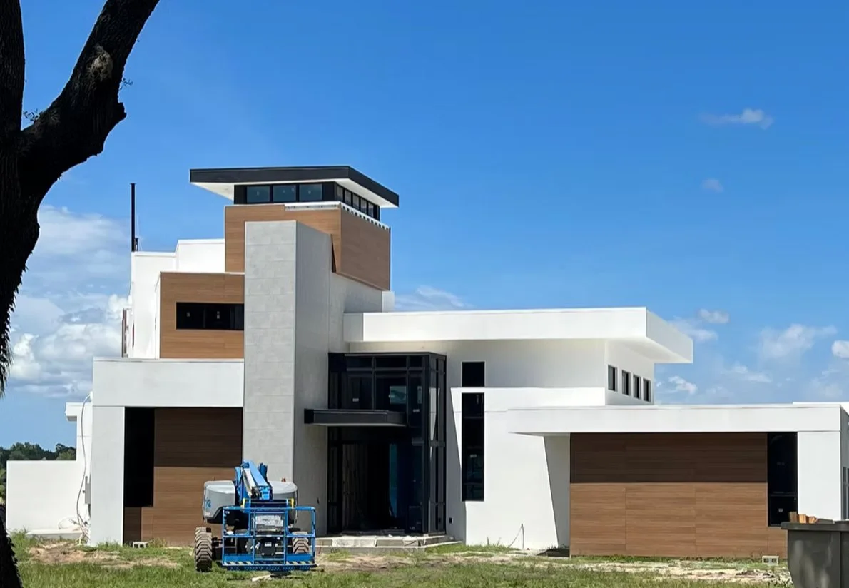 Modern multi-story house with white exterior walls, wooden accents, large windows, and a blue lift in front, under a blue sky with some clouds.