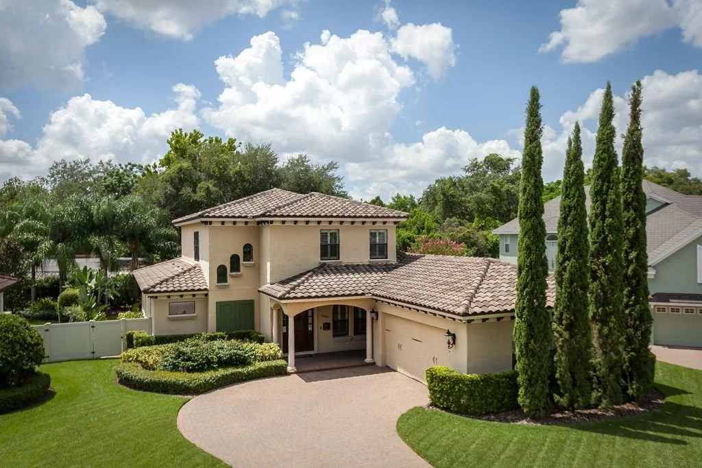 Front view of a two-story beige house with a tiled roof, a circular driveway, lush green lawn, tall cypress trees, and a partly cloudy sky.