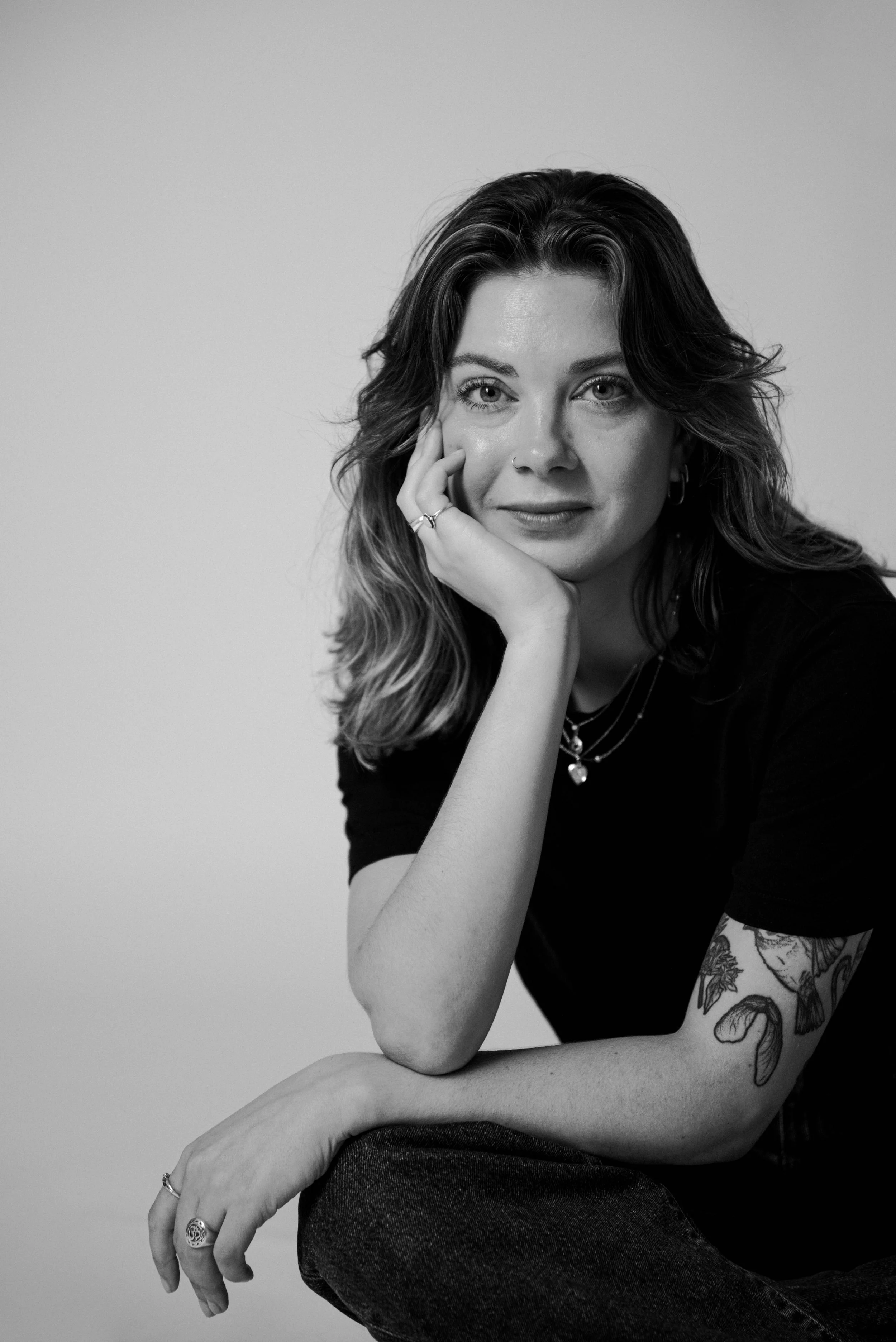 Schwarz-weiße Porträtaufnahme einer Frau mit mittellangen, gewellten Haaren, die nachdenklich wirkt, mit ihrer Hand an ihrem Gesicht, tätowiertem Arm und mit Schmuck, sitzend auf einem Stuhl.