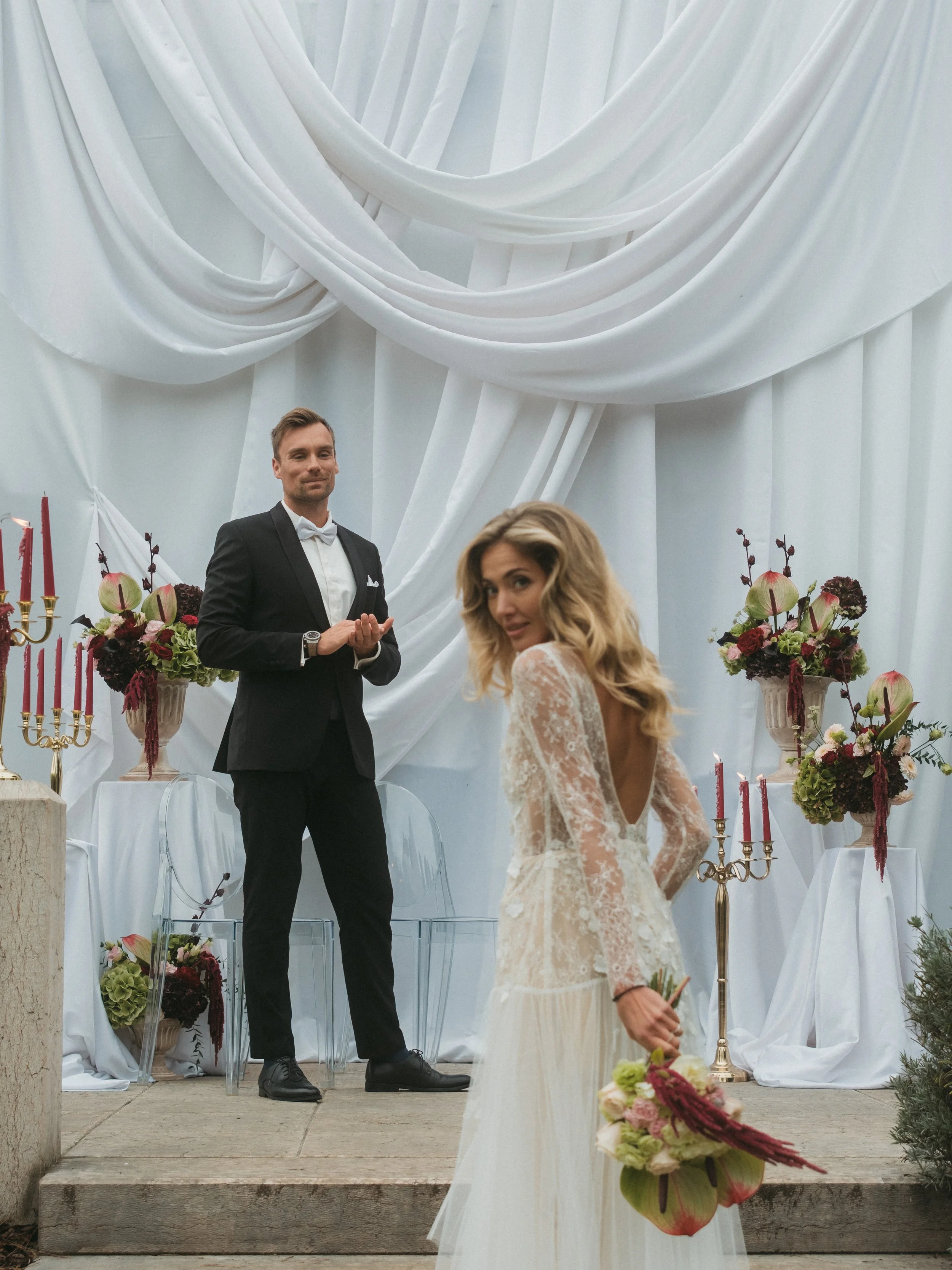 Ein Brautpaar bei einer Hochzeit, die Braut hält einen Blumenstrauß, der Bräutigam steht im Hintergrund vor einem dekorativen weißen Hintergrund mit Kerzen und Blumen.