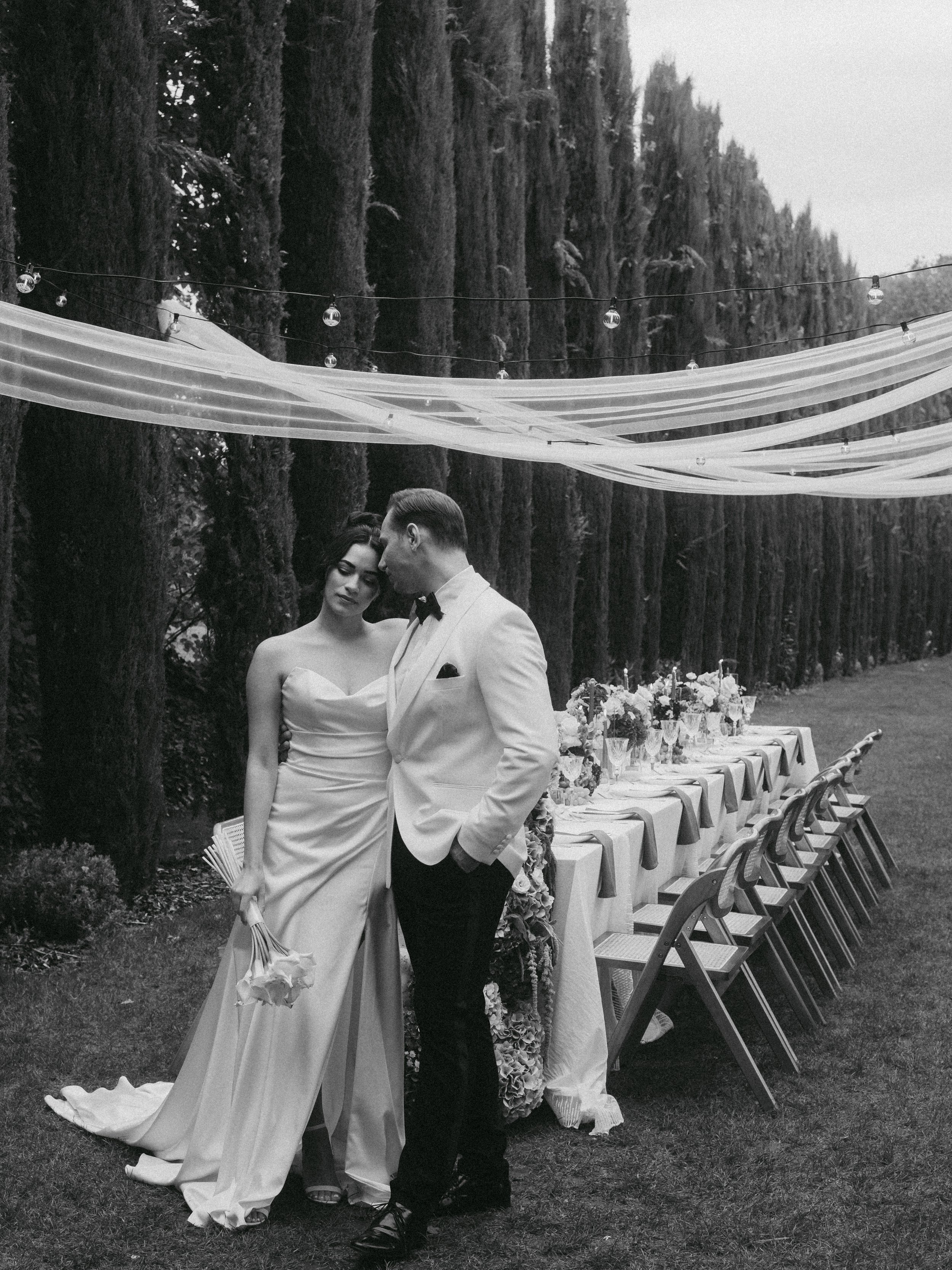 Ein Hochzeitsbild von einem Mann und einer Frau, die sich nahe stehen, im Freien mit einem langen gedeckten Tisch im Hintergrund, umgeben von hohen Bäumen und festlicher Dekoration.