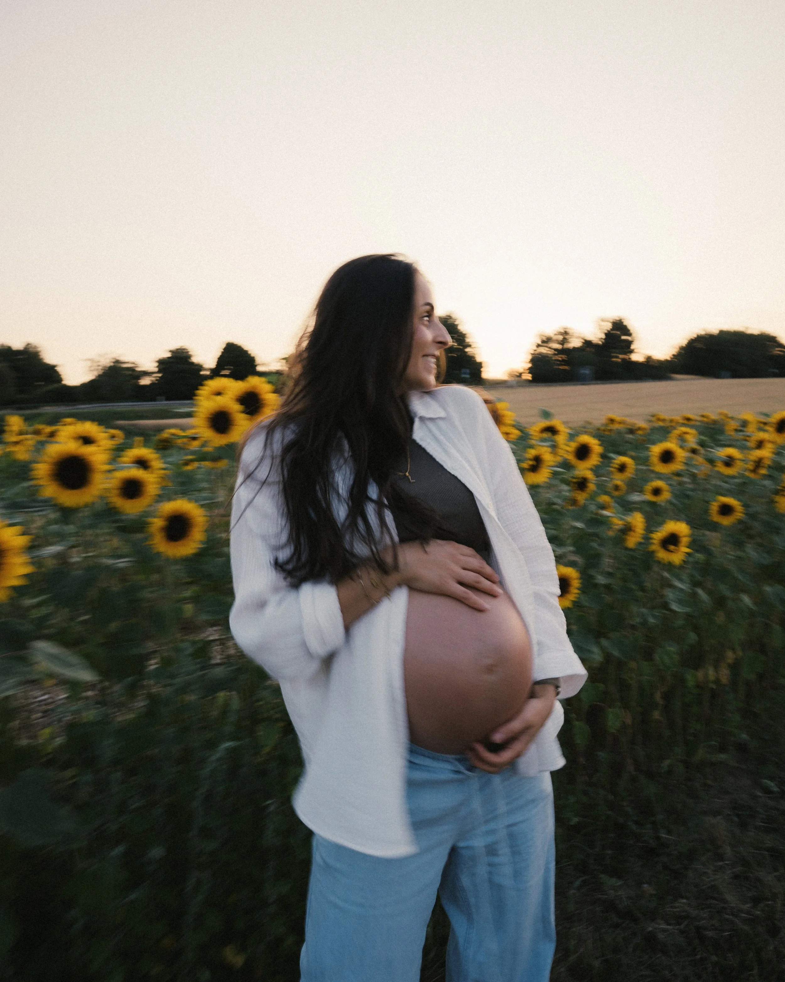 Schwangere Frau steht in einem Sonnenblumenfeld bei Sonnenuntergang und lächelt.