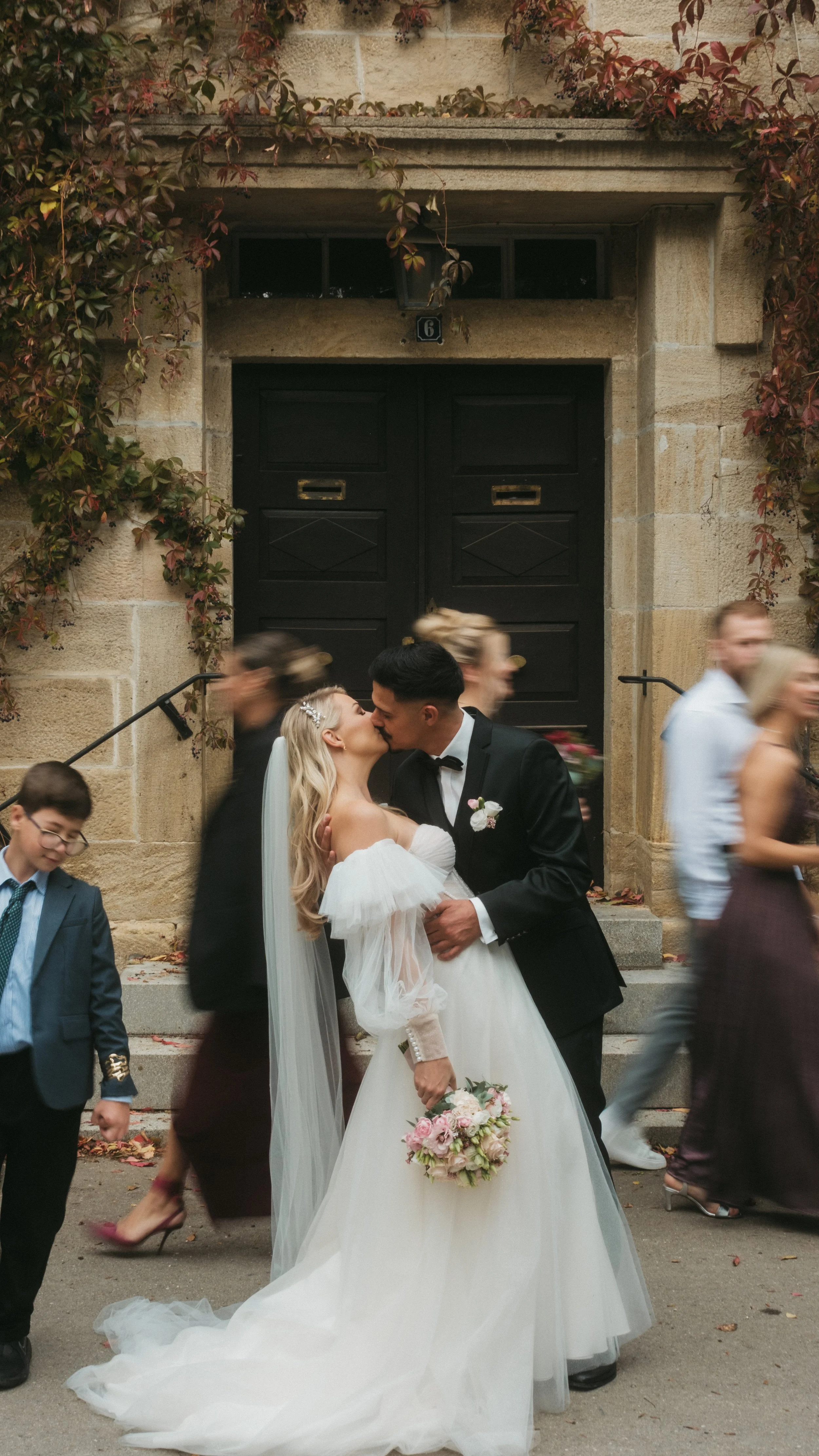 Ein frisch vermähltes Paar küsst sich vor einer alten Steintür, umgeben von fließenden Passanten bei einer Hochzeit im Freien.