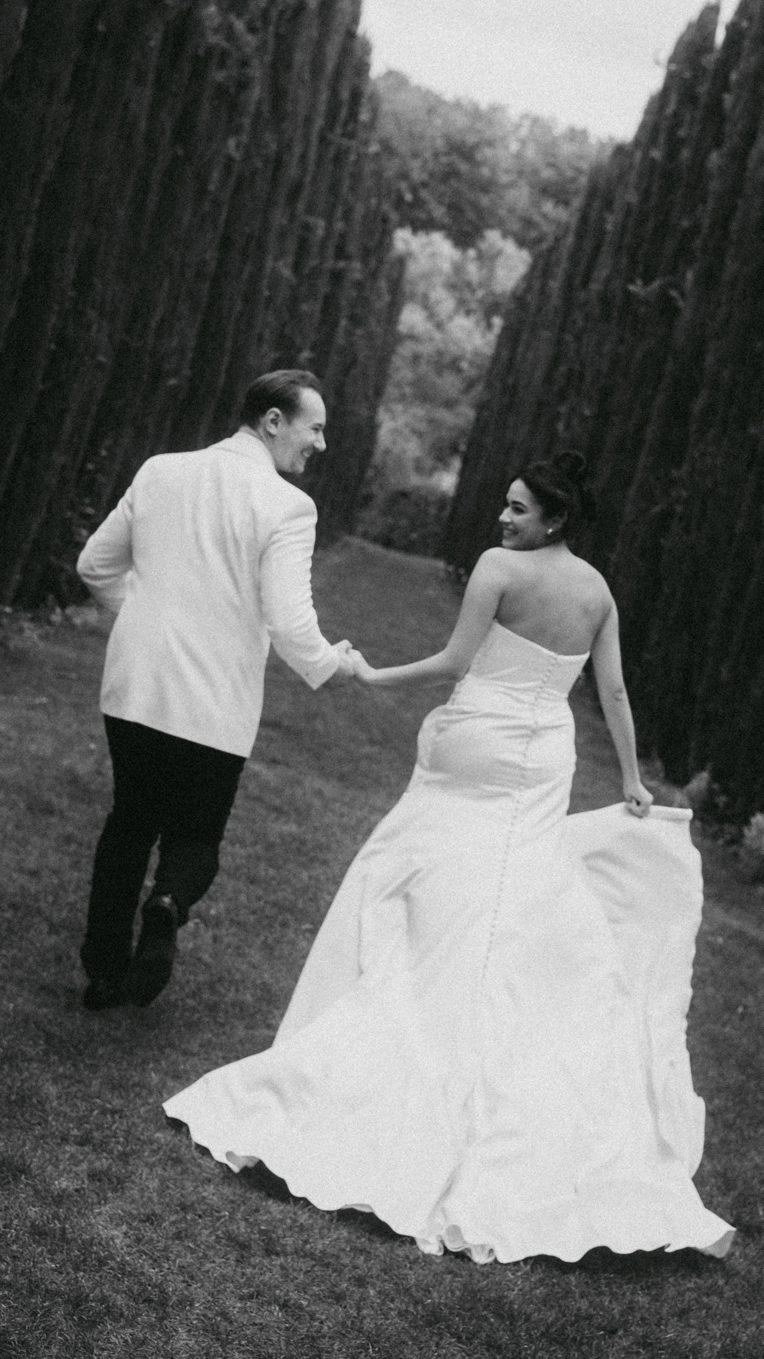 Ein glückliches Paar bei ihrer Hochzeit, tanzend im Freien zwischen hohen Baumstämmen. Die Frau trägt ein langes, weißes Brautkleid, der Mann einen weißen Anzug.