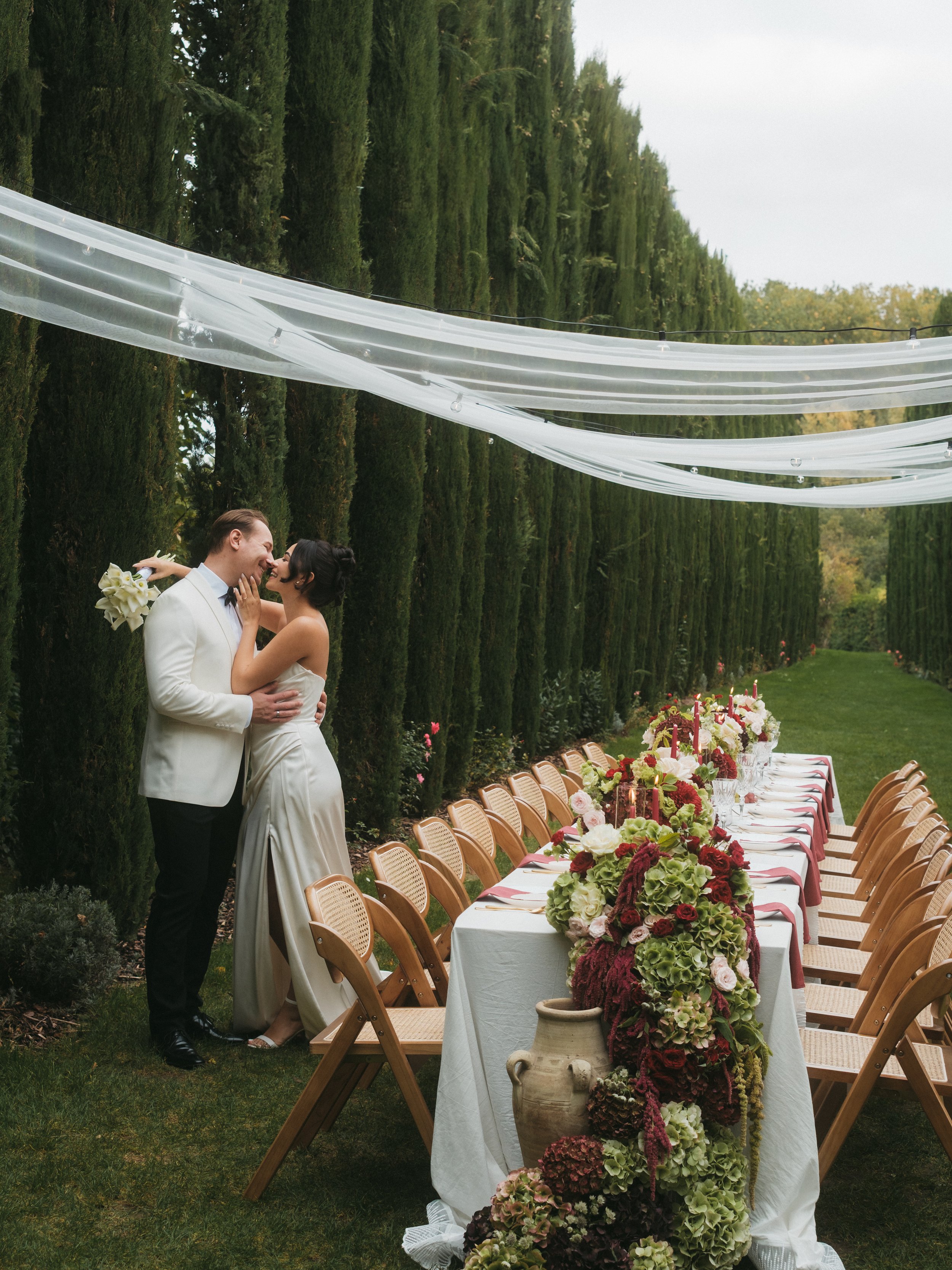 Ein Hochzeitspaar steht vor einem festlich gedeckten Esstisch im Freien mit hohen grünen Hecken im Hintergrund.