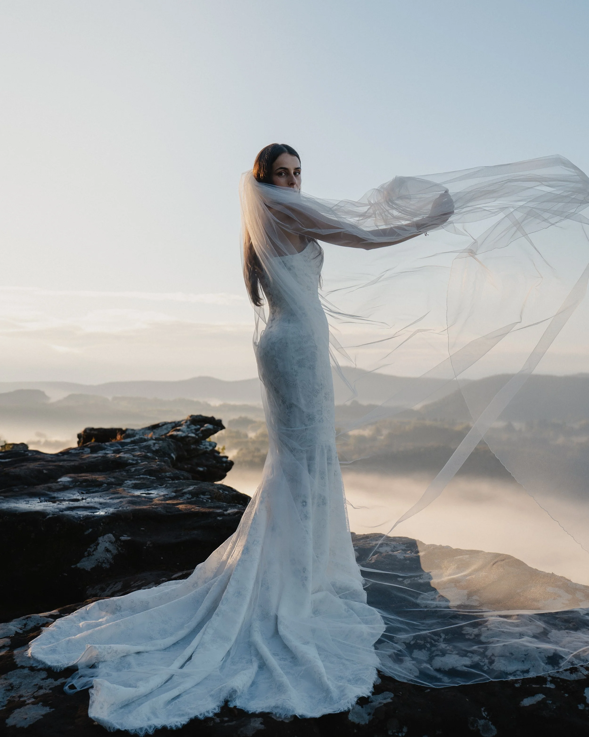 Eine Frau in einem weißen Brautkleid steht auf einem Felsen mit Blick auf eine neblige Landschaft bei Sonnenaufgang oder Sonnenuntergang und hält einen durchsichtigen Schleier, der im Wind weht.