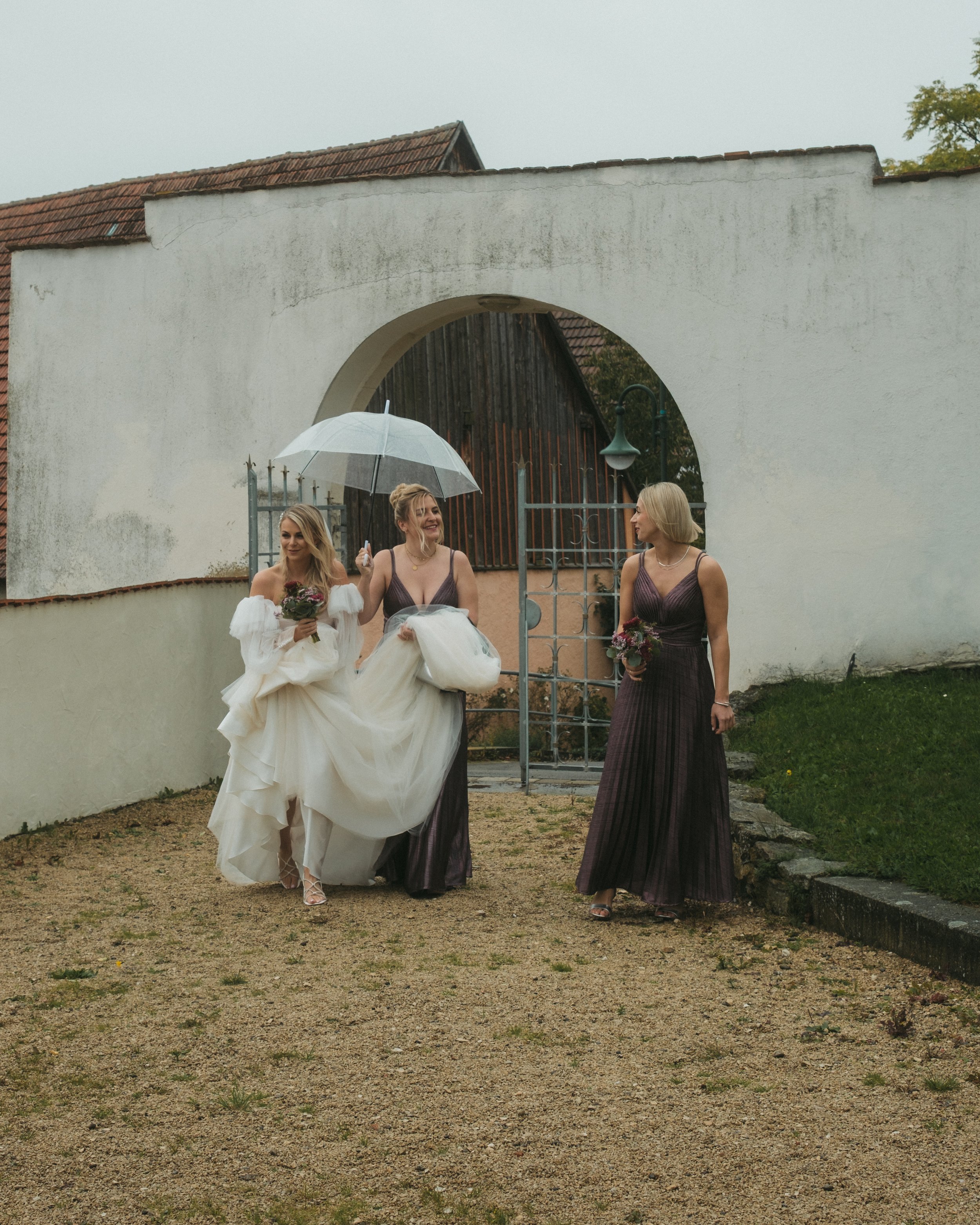 Drei Frauen in Brautkleid und Brautjungfernkleidern laufen bei Regen, eine hält einen weißen Regenschirm, alle tragen Blumensträuße, in einer historischen Toranlage und einem Garten.
