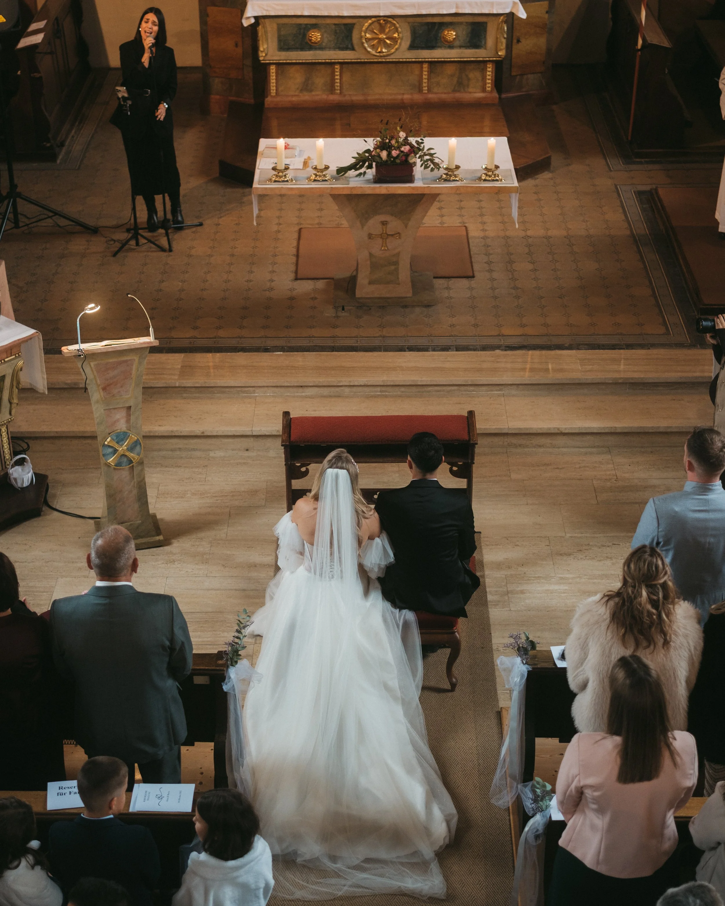 Ein Brautpaar bei einer Trauung in einer Kirche, mit Gästen und einer Sängerin am Altar.