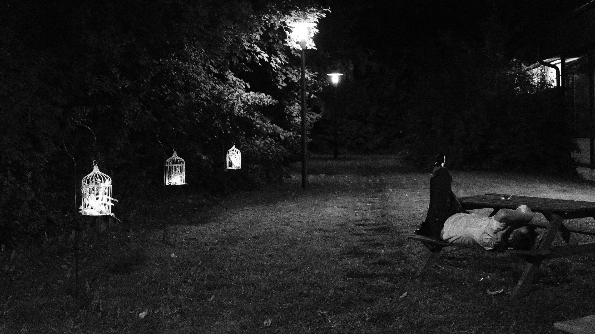 Eine nächtliche Szene im Freien mit vier beleuchteten Vogelkäfigen, einem beleuchteten Laternenpfahl und einer Person, die auf einer Bank sitzt und eine Person liegt. Das Bild ist in Schwarzweiß.