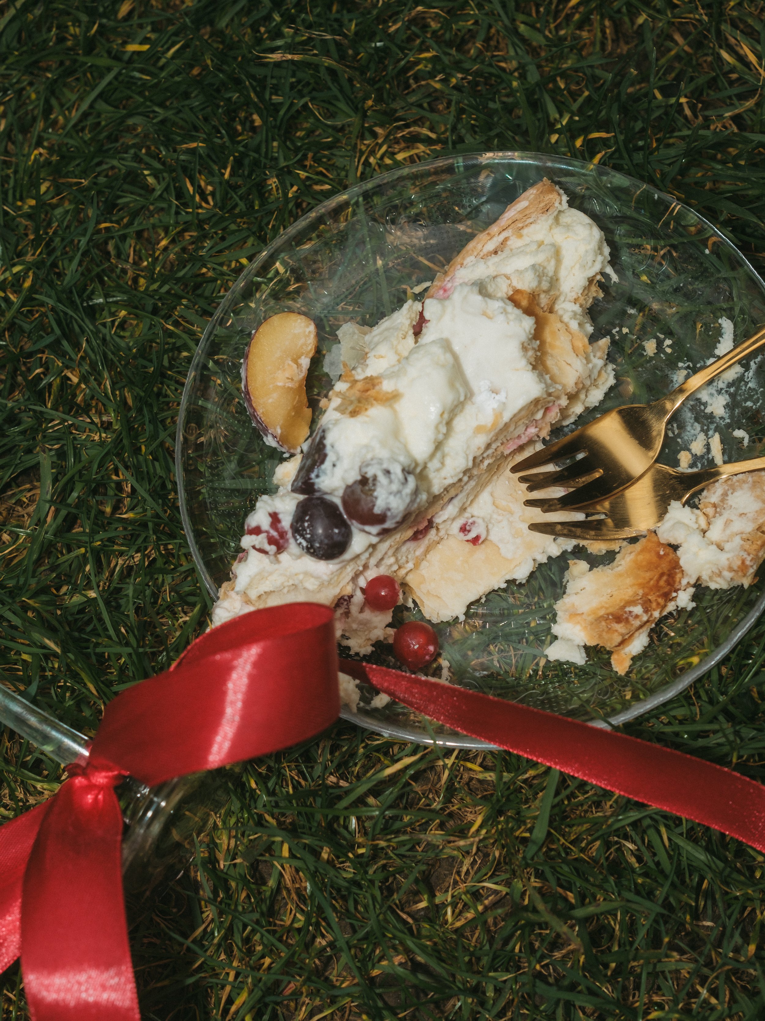 Verzehrtes Stück Torte mit Beeren und einem Löffel auf einem Glasdessertteller, dekoriert mit einem roten Band, liegt auf grünem Gras.