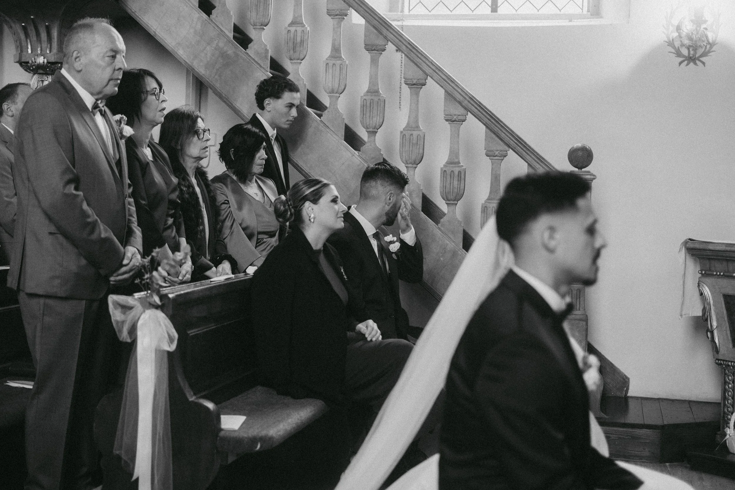 Menschen bei einer Trauung in einer Kirche, einige sitzen, andere stehen, alle zeigen einen emotionalen Ausdruck, schwarz-weiß Fotografie.