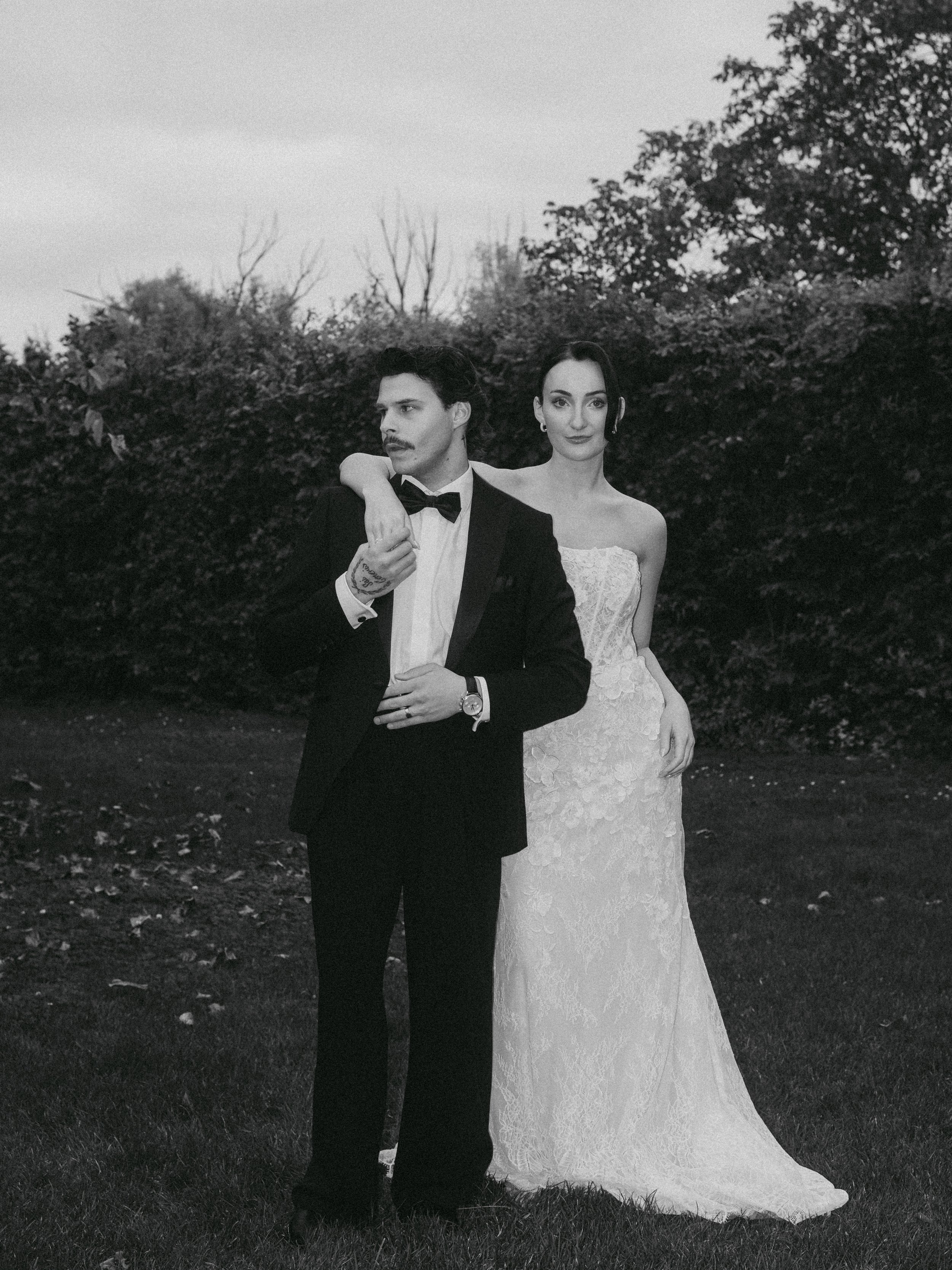 Ein elegantes Paar in formeller Kleidung steht draußen vor einer Baumhecke, der Mann trägt einen schwarzen Anzug mit Fliege, die Frau trägt ein langes, viktorianisch inspiriertes Hochzeitskleid.