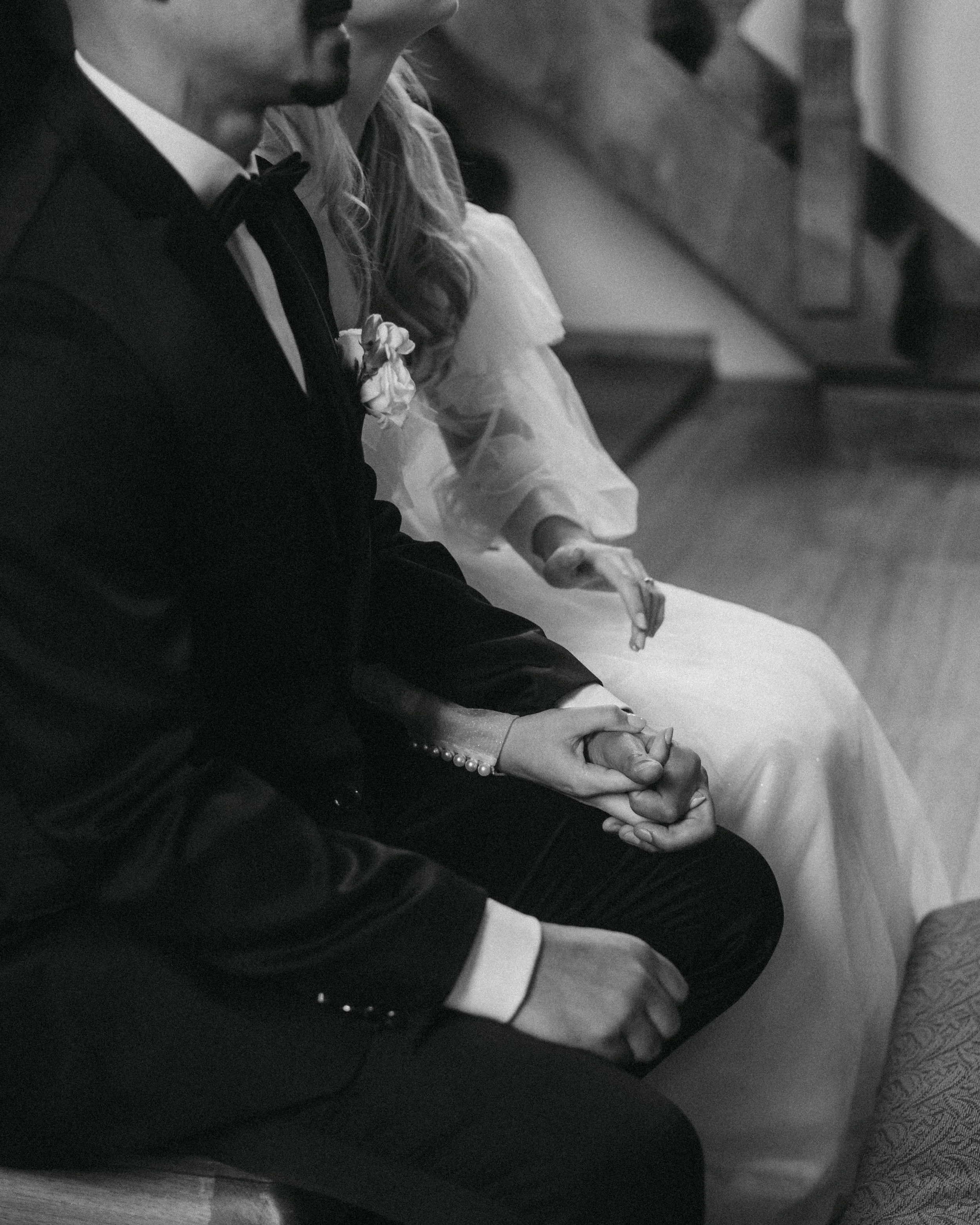 Ein Brautpaar hält sich während einer Hochzeitszeremonie an den Händen. Das Bild ist schwarz-weiß und zeigt den oberen Körper des Paares, das auf einer Bank sitzt.