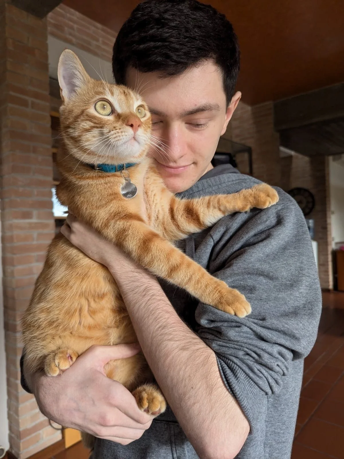 Young man holding an orange tabby cat indoors, both looking outside with the man smiling softly.