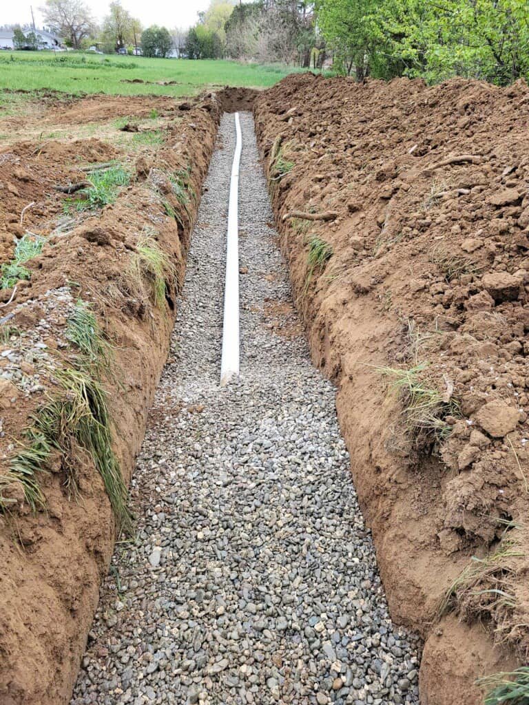 A freshly dug trench by Robert Burt Construction with gravel and a white pipe installed, likely for utility extension or drainage, with grassy field and trees in the background.