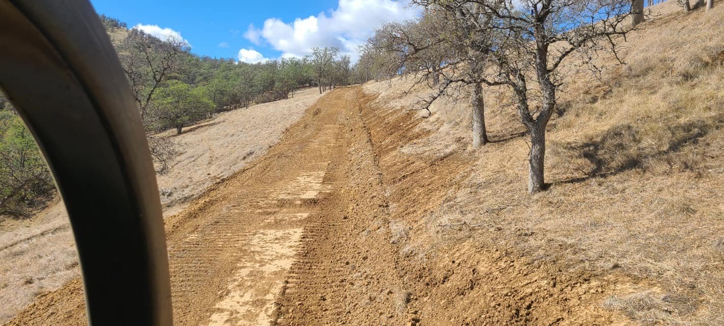 A dirt road built by Robert Burt Construction, on a hillside with trees and grass, partly cloudy sky in the background.