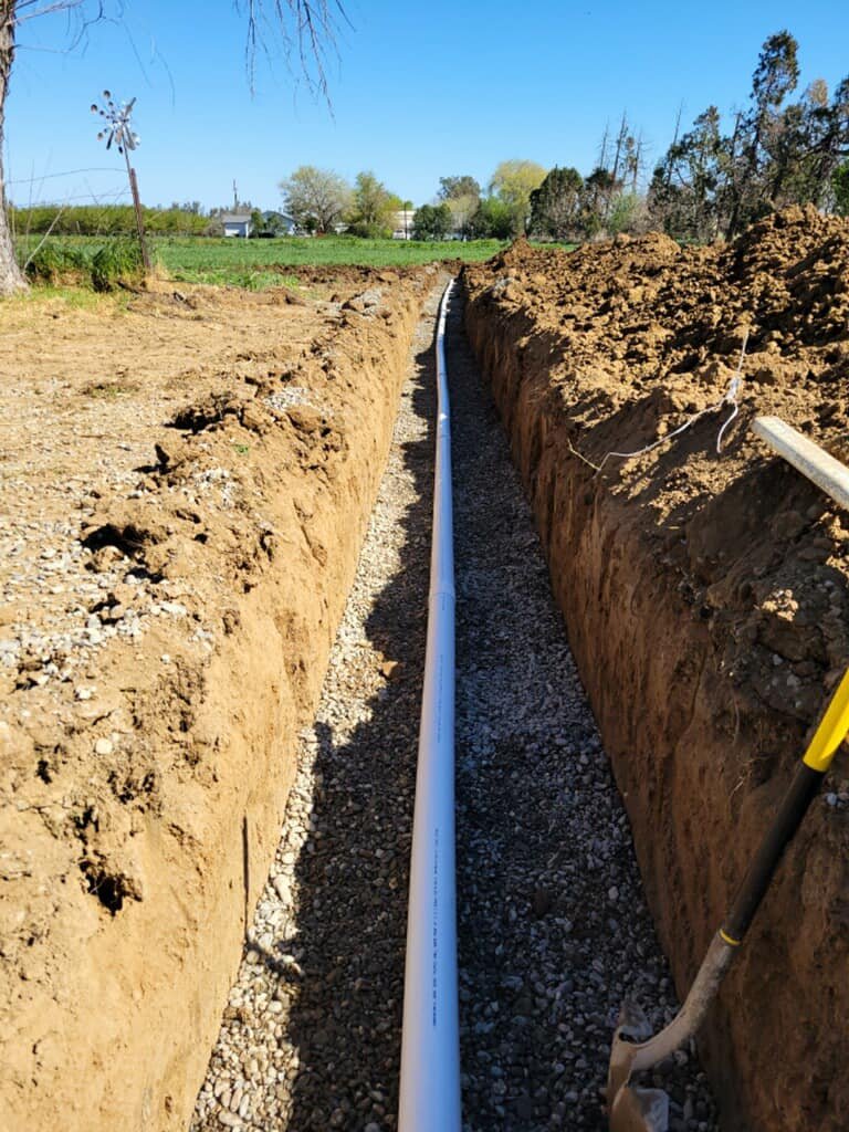 A trench with a white PVC pipe installed, view of a Robert Burt Construction site outdoors with trees and grass in the background.