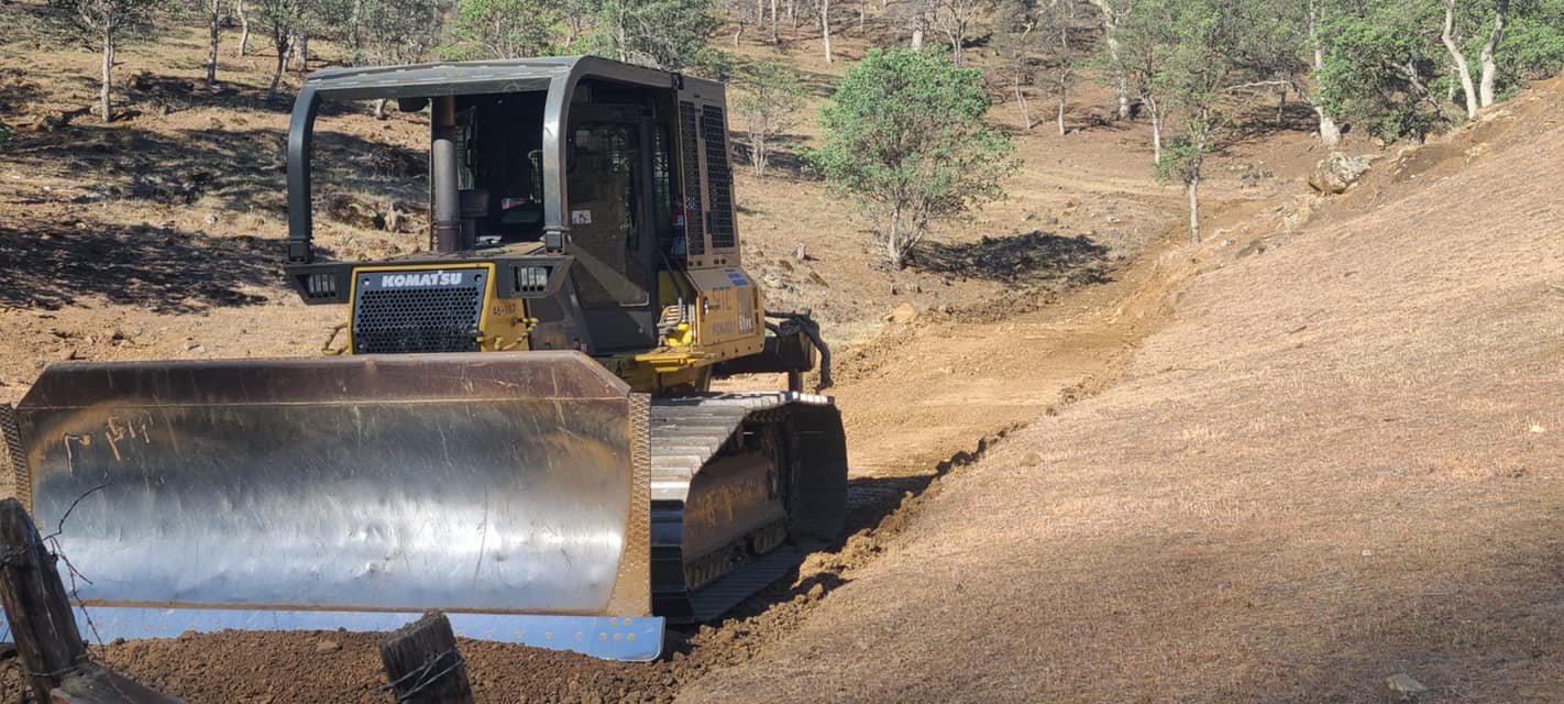 A small Robert Burt Construction Komatsu bulldozer leveling the ground with a large metal blade in a dirt construction site surrounded by trees.