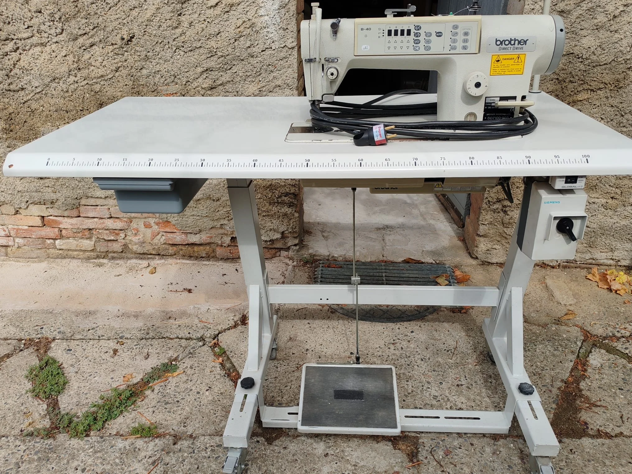 Machine à coudre industrielle Brother montée sur une table blanche, avec un panneau de contrôle, située à l'extérieur sur un sol en béton avec des feuilles mortes et une brique en arrière-plan.