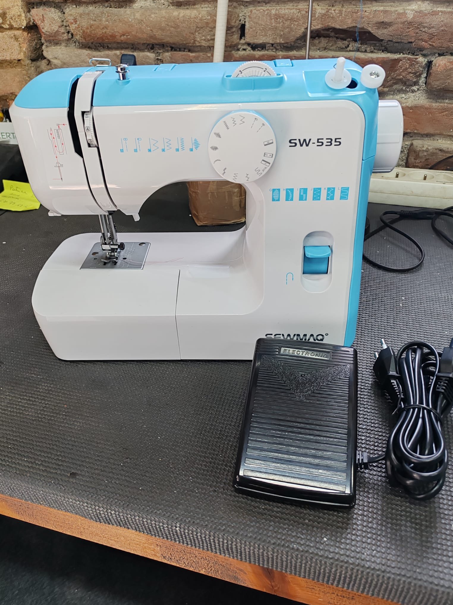 Une machine à coudre blanche et bleue sur une table en bois, avec un pied presseur et divers réglages, à côté d'une pédale, dans un environnement industriel ou de couture.