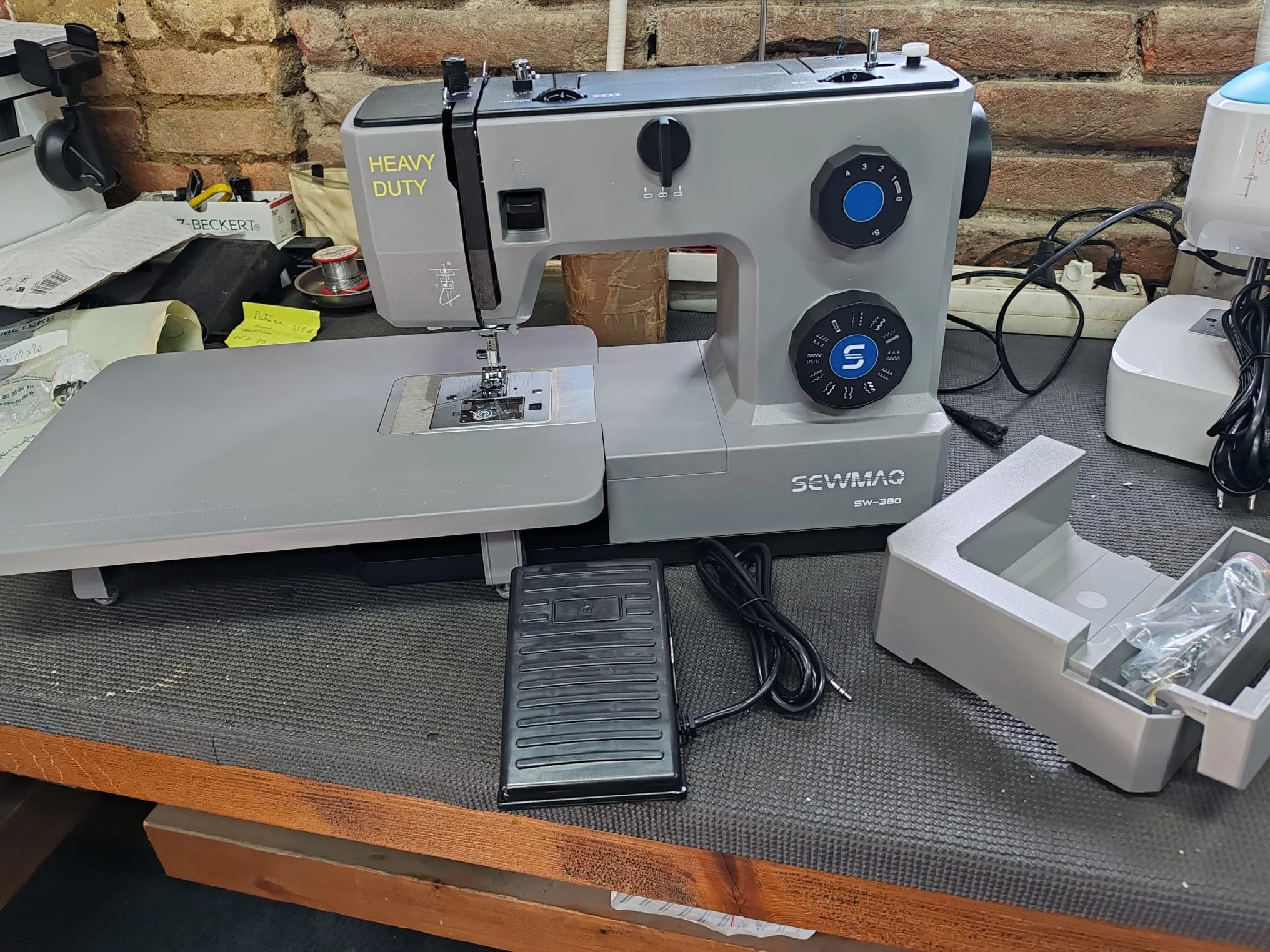 Une machine à coudre de marque Sewmaq sur une table avec divers outils et matériaux autour, contre un mur de briques.
