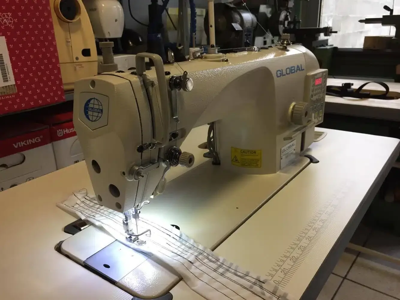 Une machine à coudre industrielle blanche sur une table, avec un tissu rayé en dessous du pied-de-boulon, dans un atelier de couture.