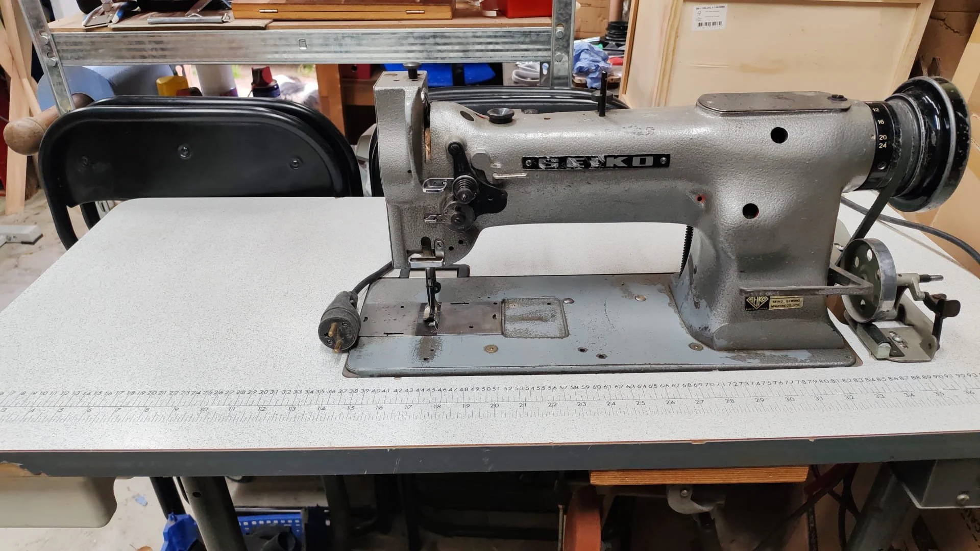 Une vieille machine à coudre en métal sur une table de travail, avec une règle de mesure en dessous, dans un atelier ou un espace de bricolage.
