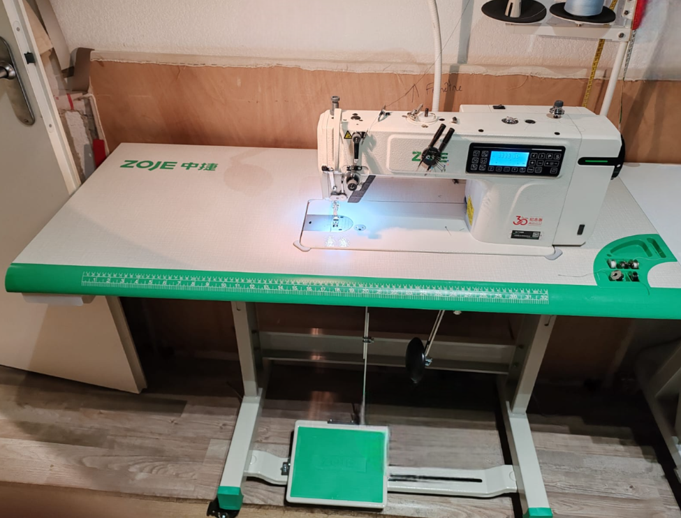 Une machine à coudre blanche sur une table de couture, avec un guide de mesure vert, située dans un atelier de couture.