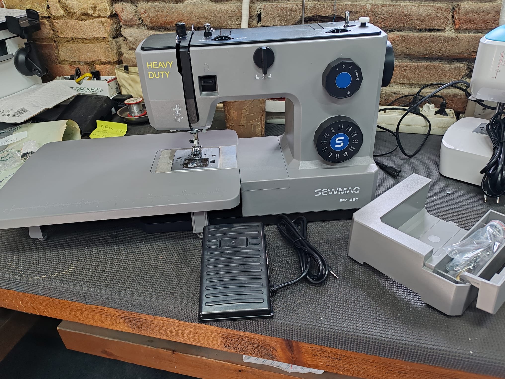 Une machine à couture de marque Sewmaq sur une table en bois, entourée de divers outils et matériaux, avec un mur en briques en arrière-plan.