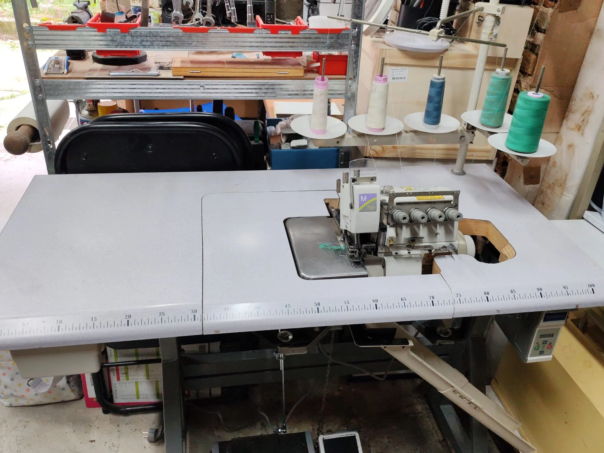 Une machine à coudre industrielle sur une table de travail avec plusieurs bobines de fil suspendues à l'arrière, dans un atelier de couture ou de fabrication textile.