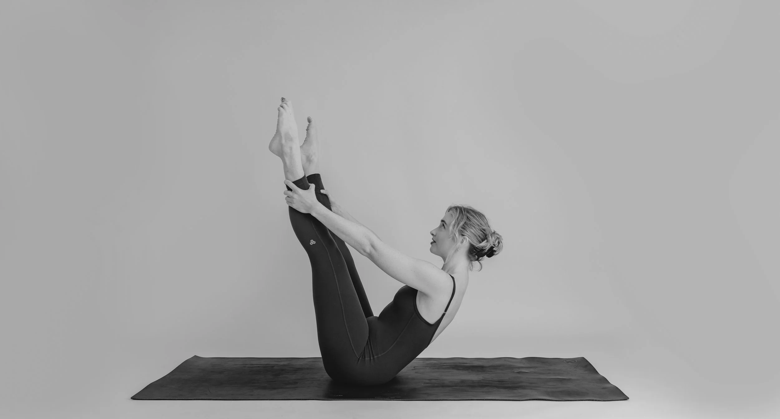 A woman practicing Pilates in a black tank top and leggings. She is performing the Pilates pose, The Open Leg Rocker.