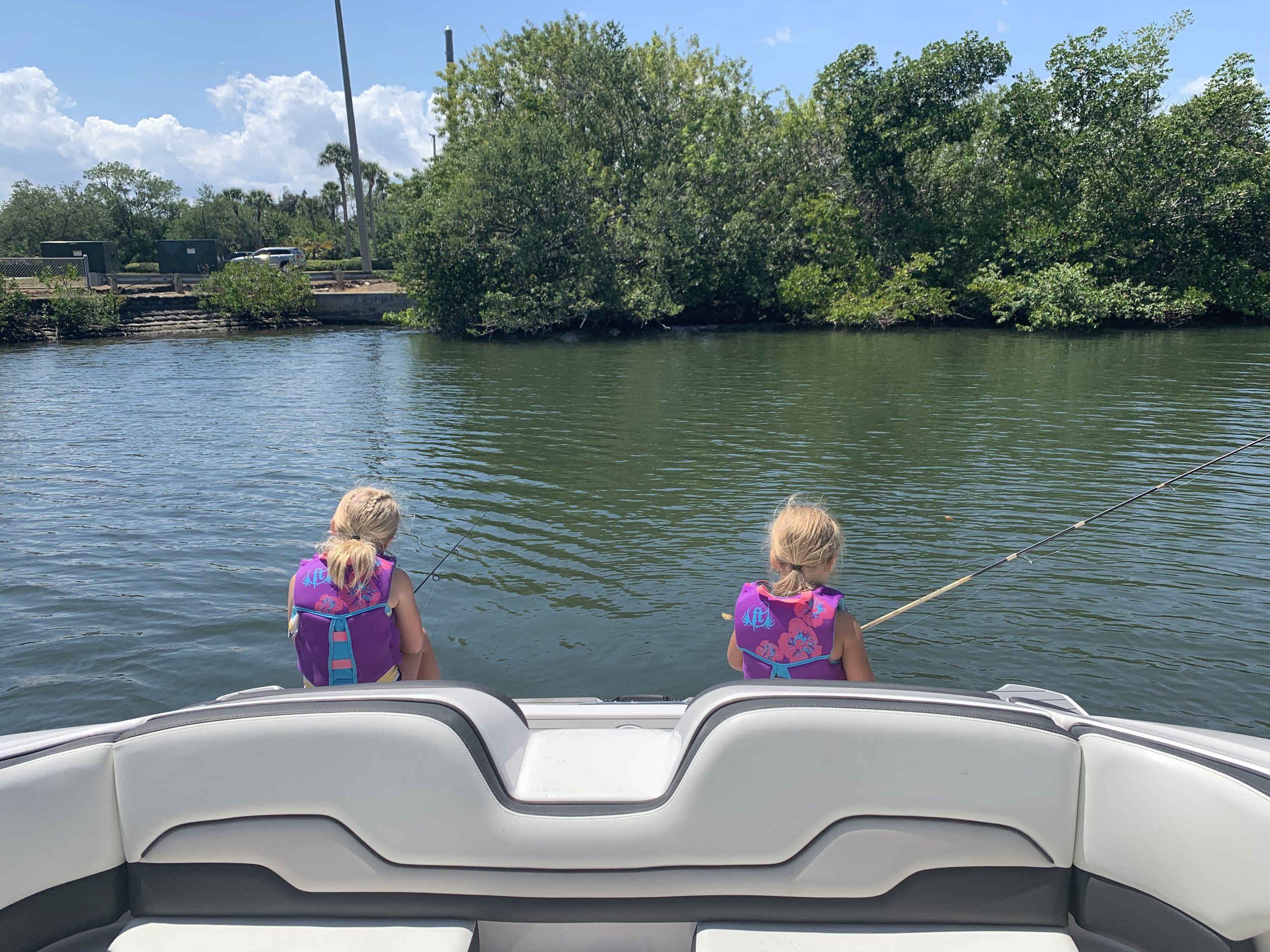 Two young girls wearing purple life jackets in a boat, fishing in a calm river with green trees and a partly cloudy sky in the background.