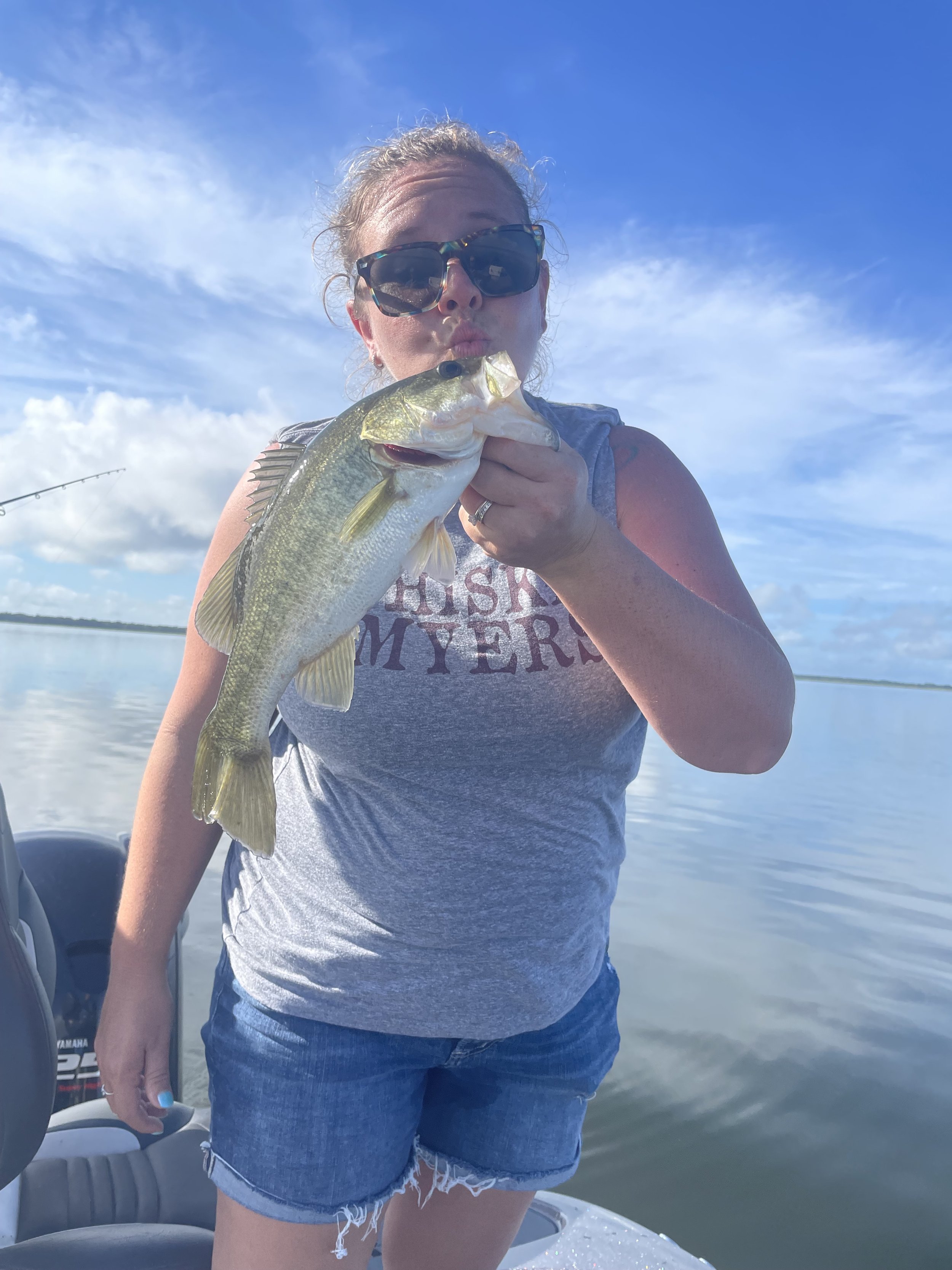 Woman wearing sunglasses and a gray tank top holding a largemouth bass fish on a boat in a water body under a blue sky with clouds.