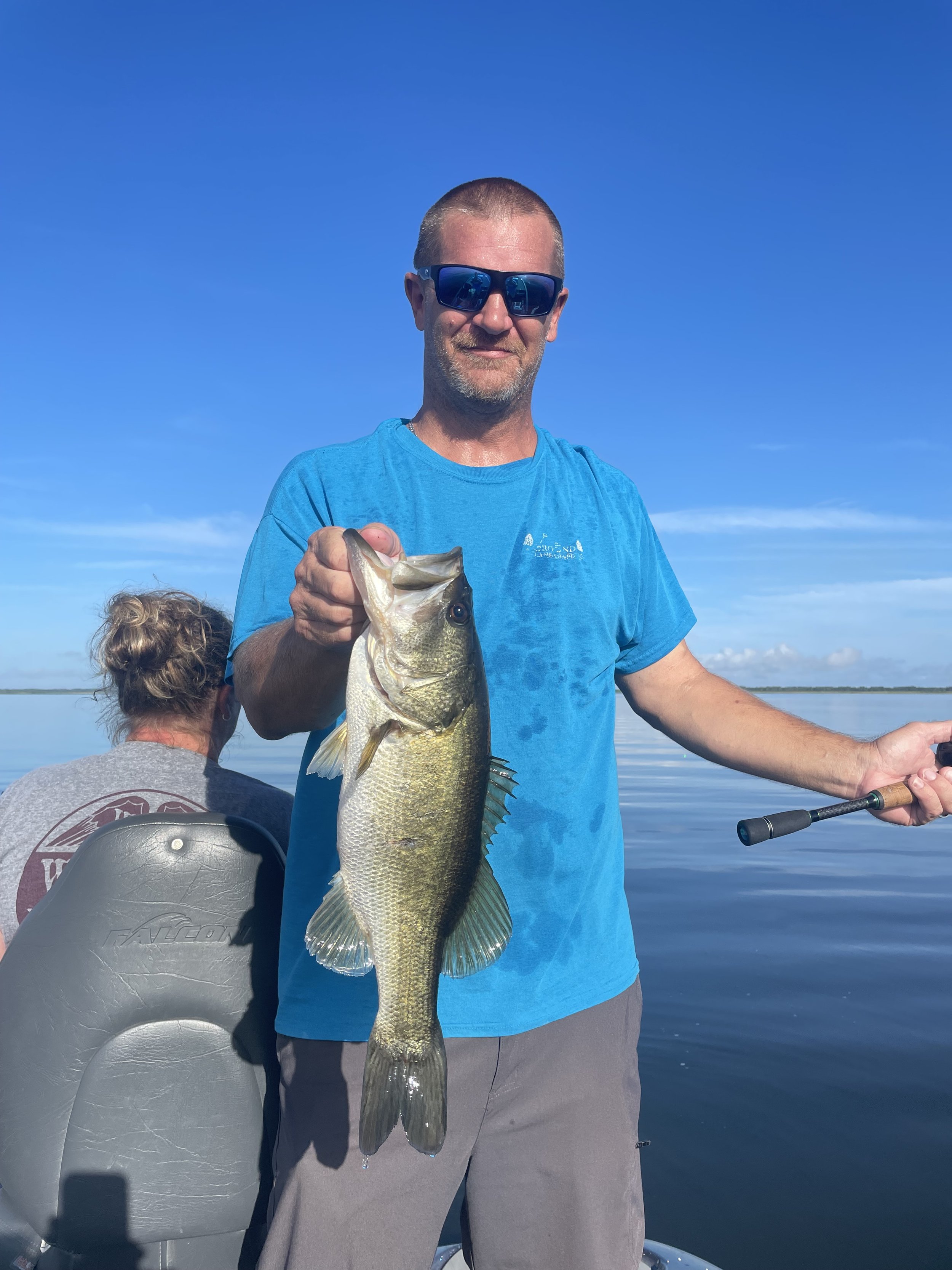 Man wearing sunglasses holding a large fish on a boat in a calm lake with another person sitting behind him, and a clear blue sky in the background.