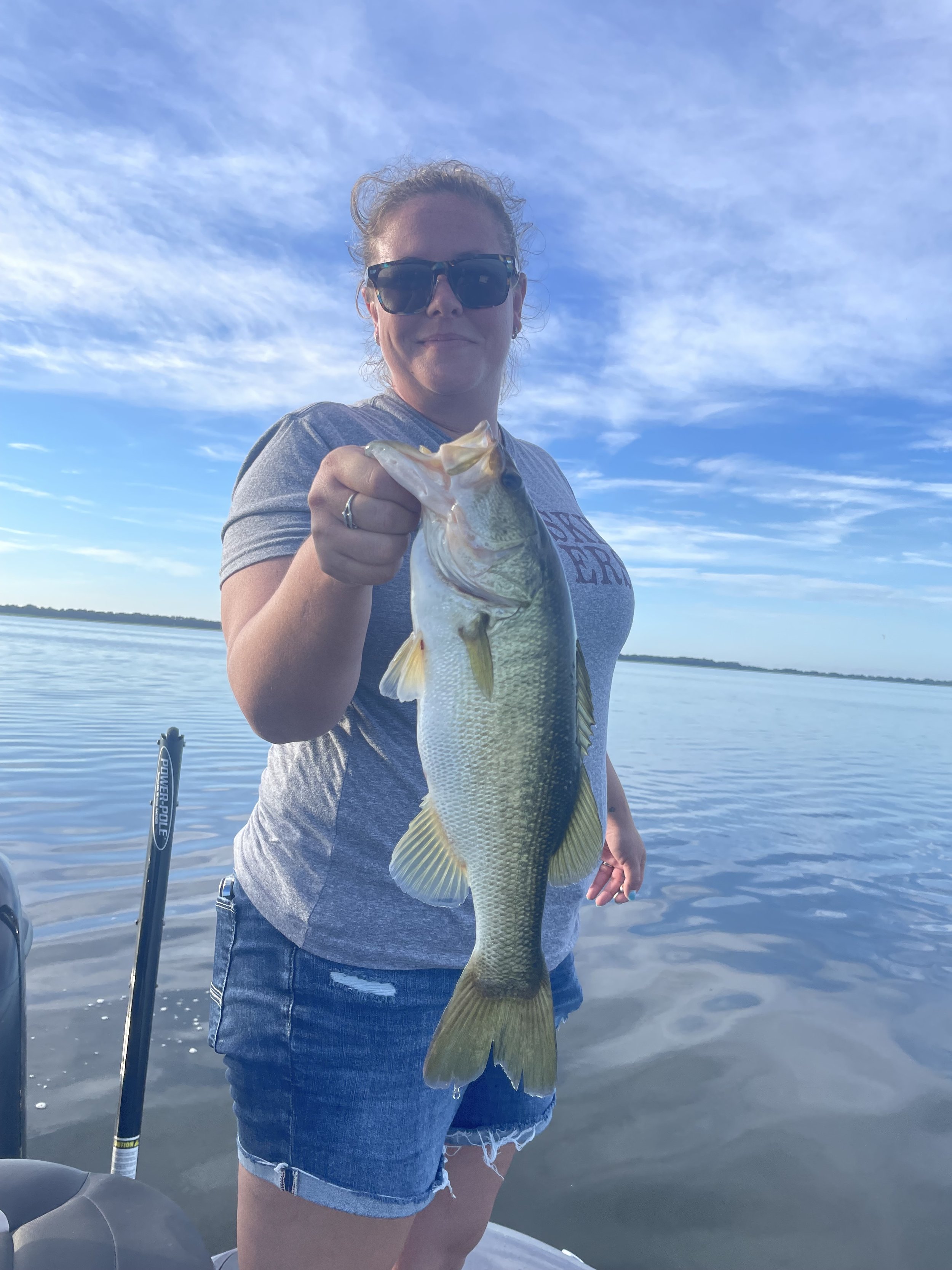 Woman in sunglasses holding a large bass fish on a boat in a calm lake with a blue sky and clouds in the background.