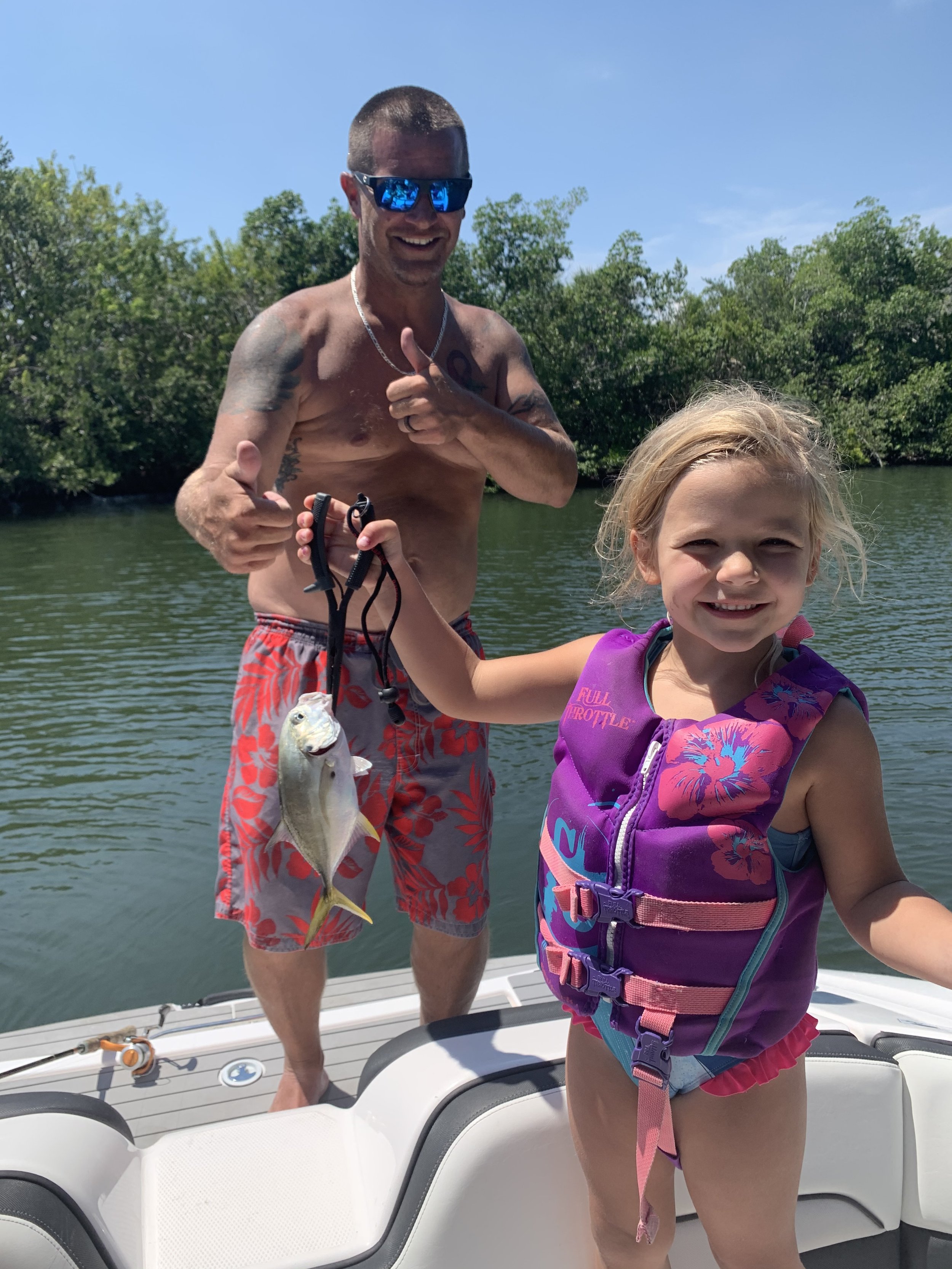 A man and a young girl on a boat after fishing. The man is shirtless, wearing sunglasses, floral shorts, and smiling with a thumbs-up. The girl is holding a fish, wearing a purple life jacket, smiling, with water and trees in the background.