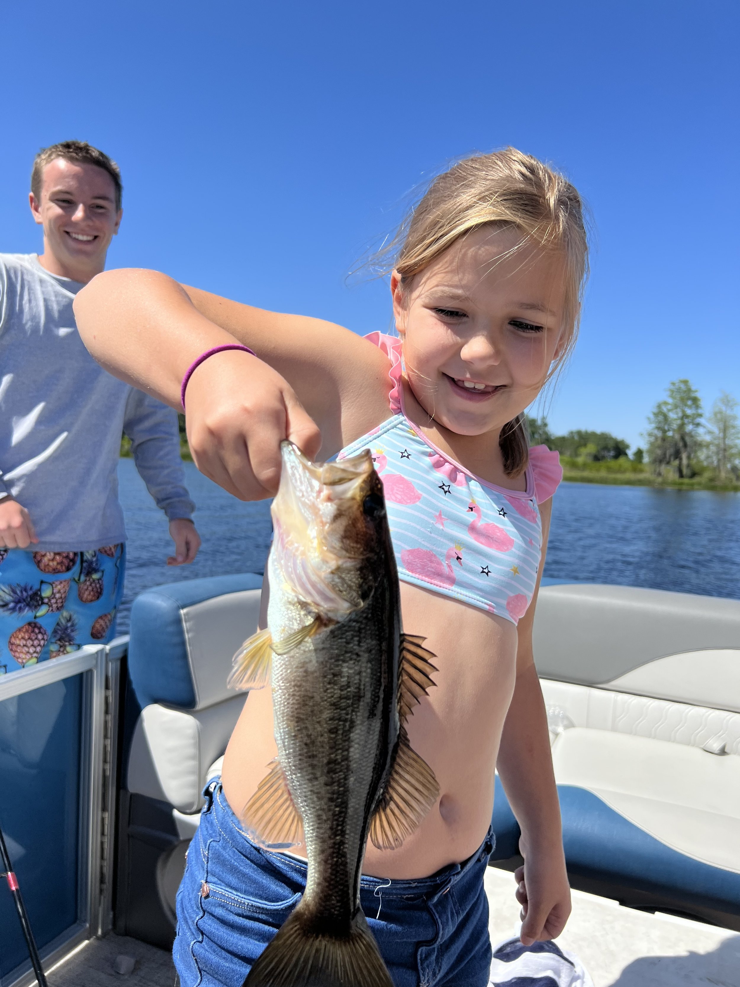 A young girl on a boat holding a fish she caught, smiling, with a man in the background near a body of water on a sunny day.