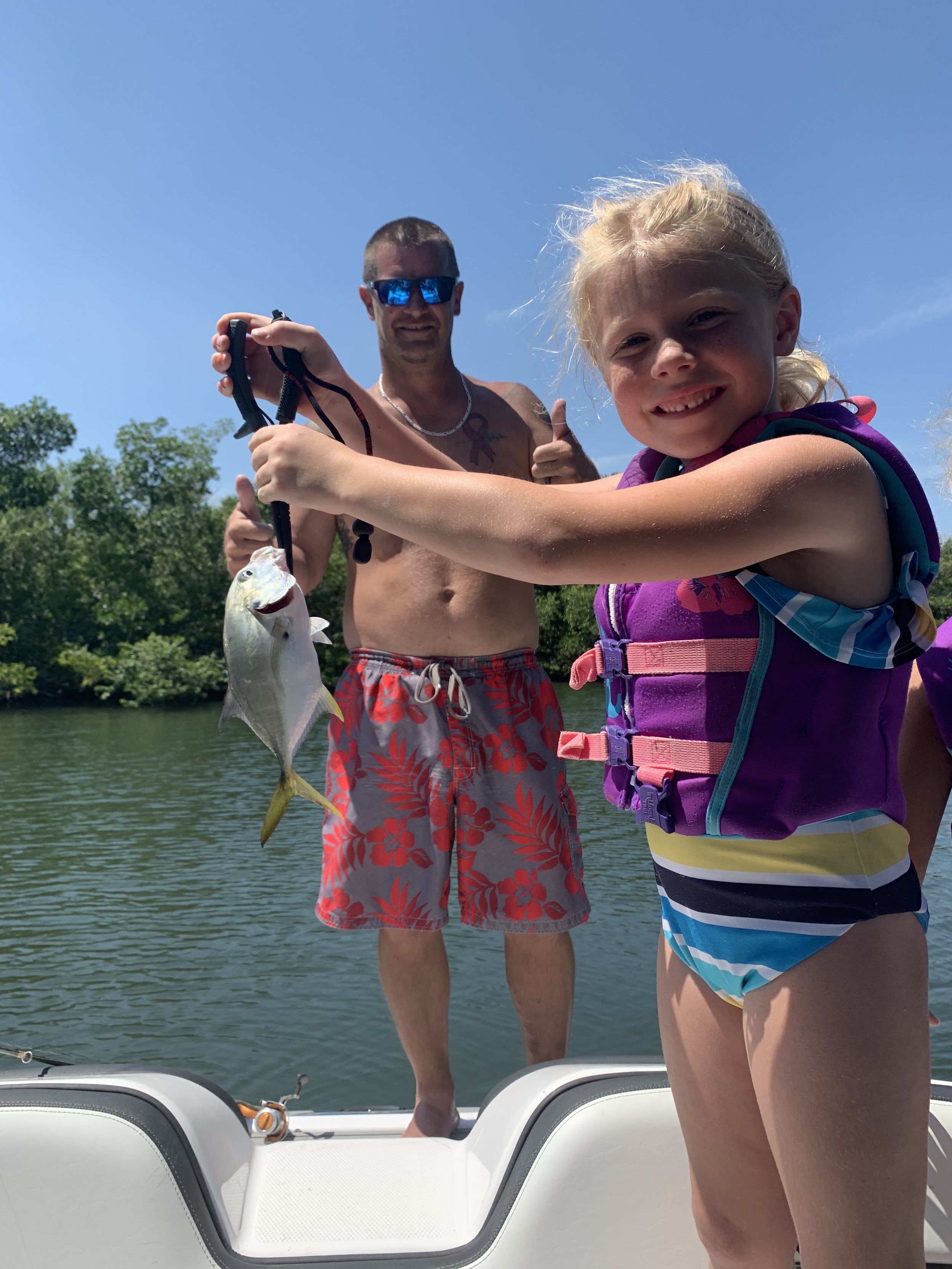 A young girl in a purple life jacket holding up a fish she caught, with a smiling man in sunglasses and swim trunks behind her on a boat, giving a thumbs up. The background shows water and trees under a clear blue sky.