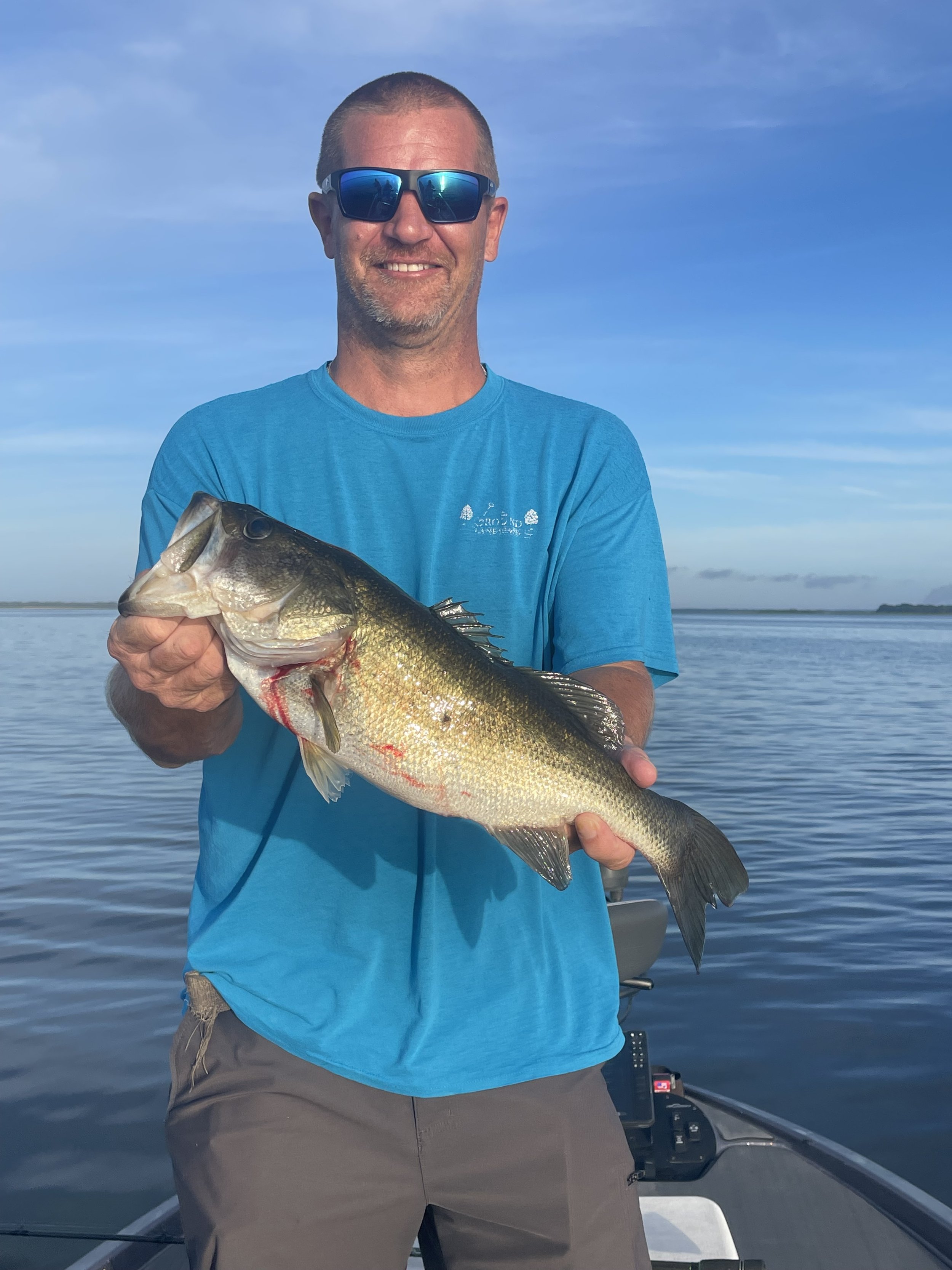 Man in blue t-shirt and sunglasses holding a large fish on a boat on a calm water body with a distant shoreline and blue sky in the background.
