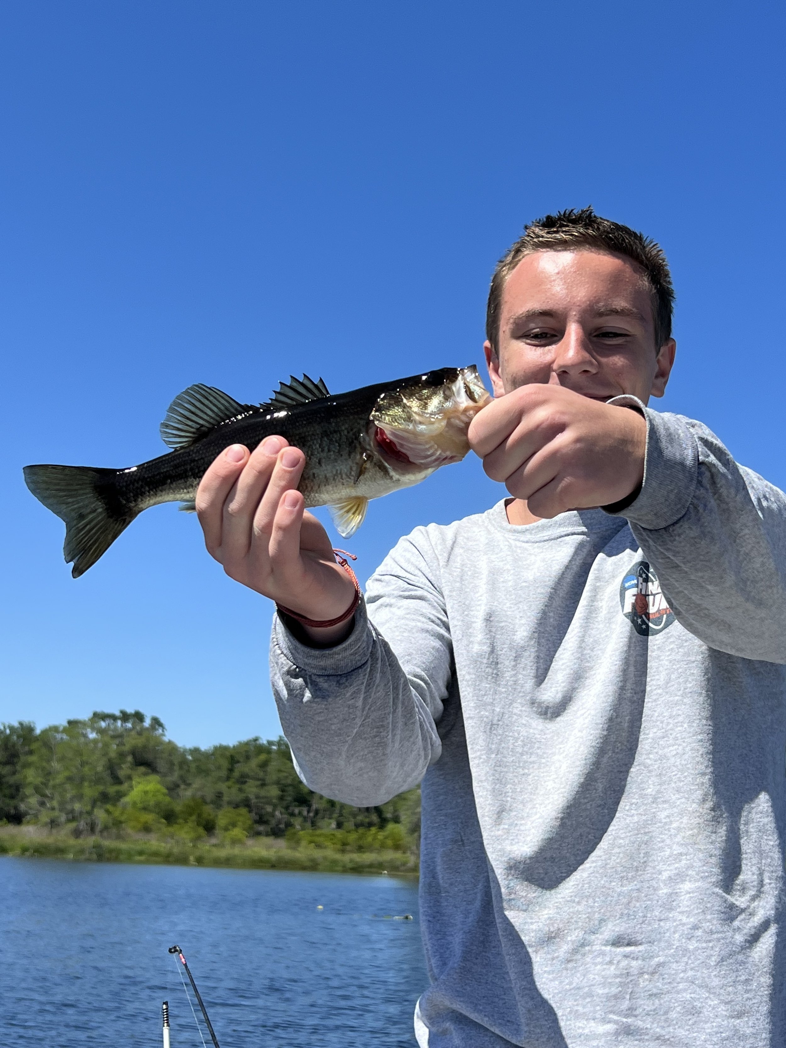 Young man holding a large fish in his right hand outdoors by a lake and blue sky.