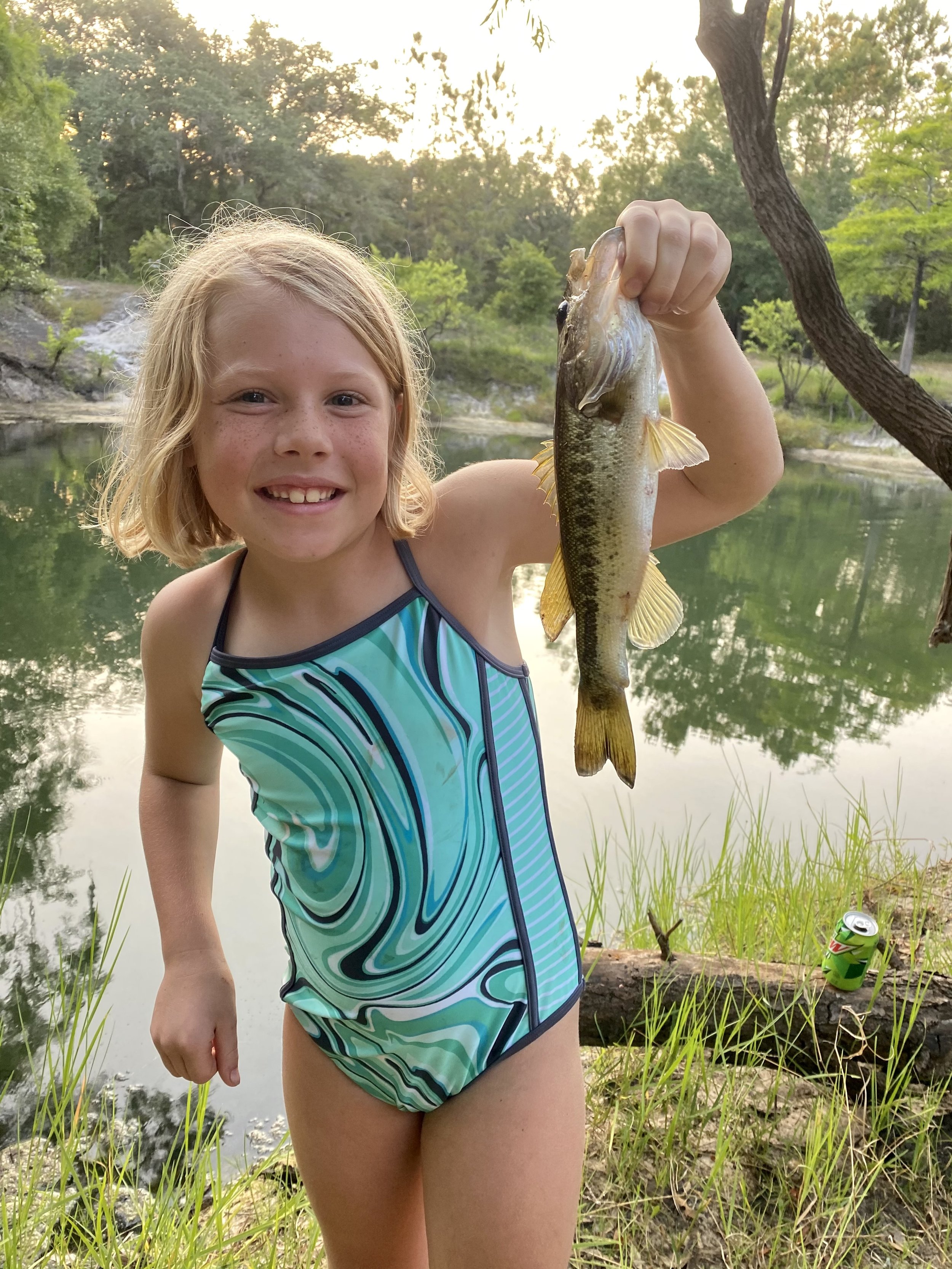 A young girl with blonde hair smiling and holding a fish she caught, standing near a pond with trees and a soda can in the background.