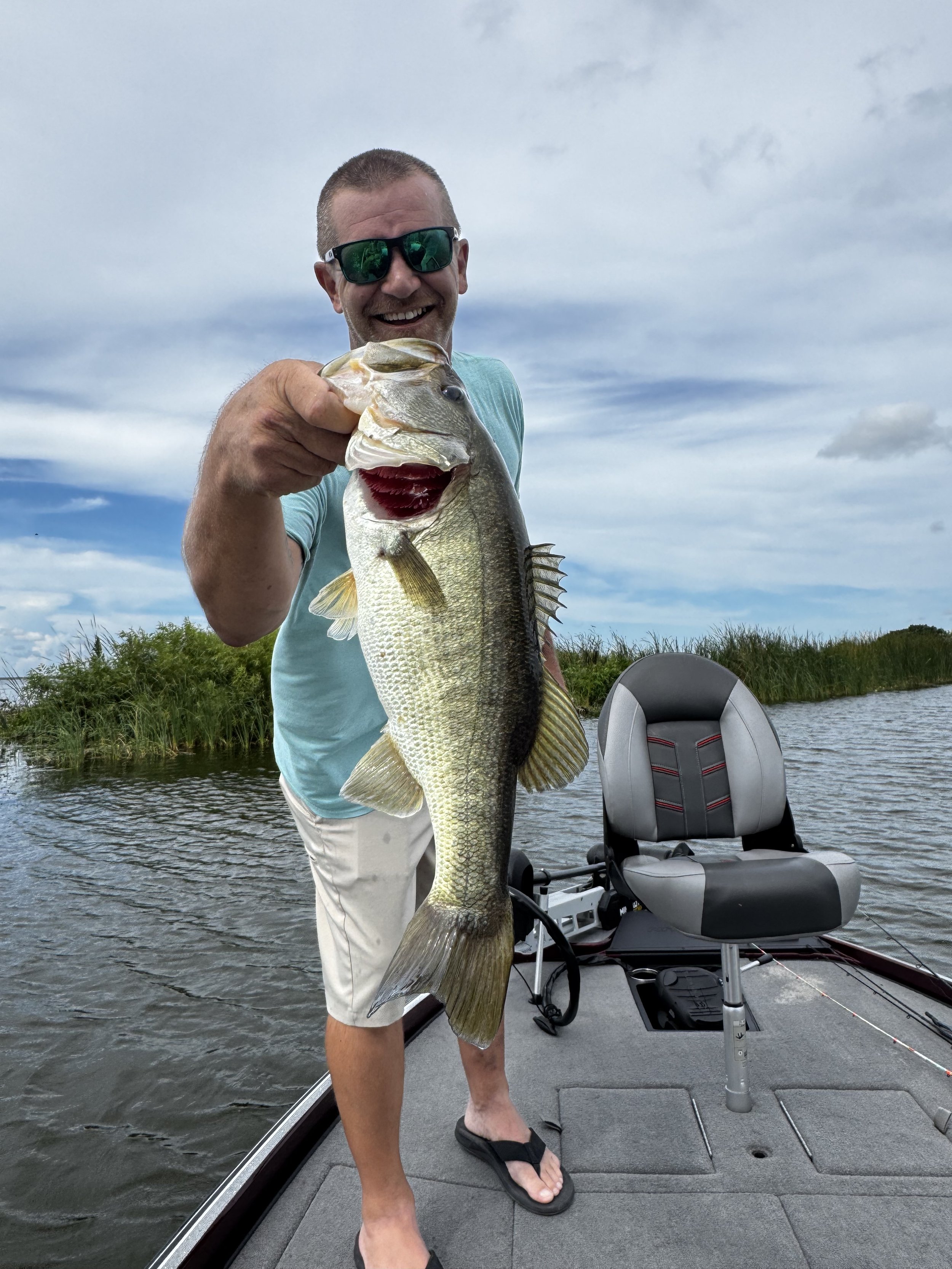 Man in sunglasses holding a large bass fish on a boat in a lake, with water and reeds in the background.