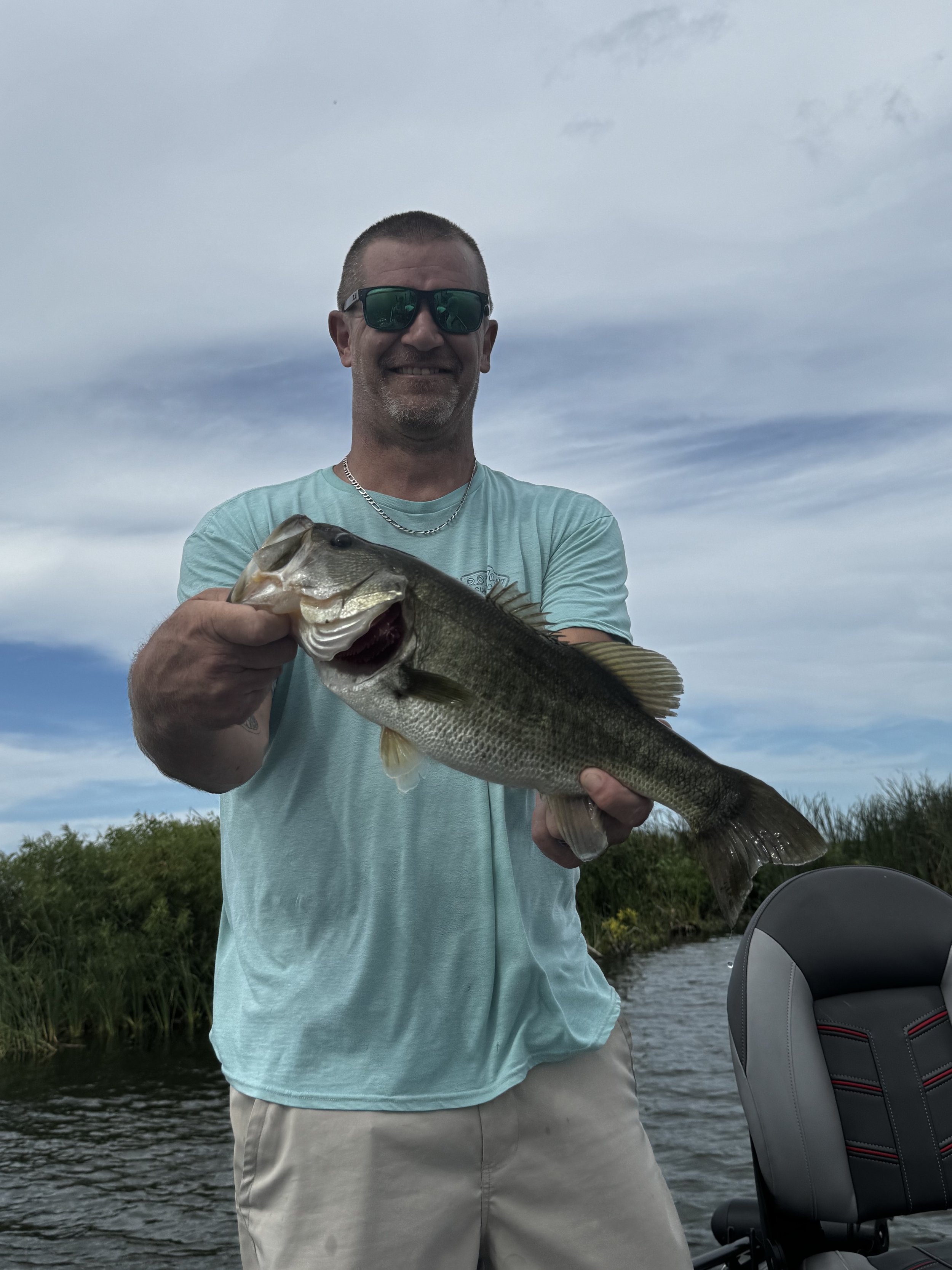 A man wearing sunglasses and a light blue t-shirt holding a largemouth bass fish outdoors near a body of water with grassy shoreline in the background.