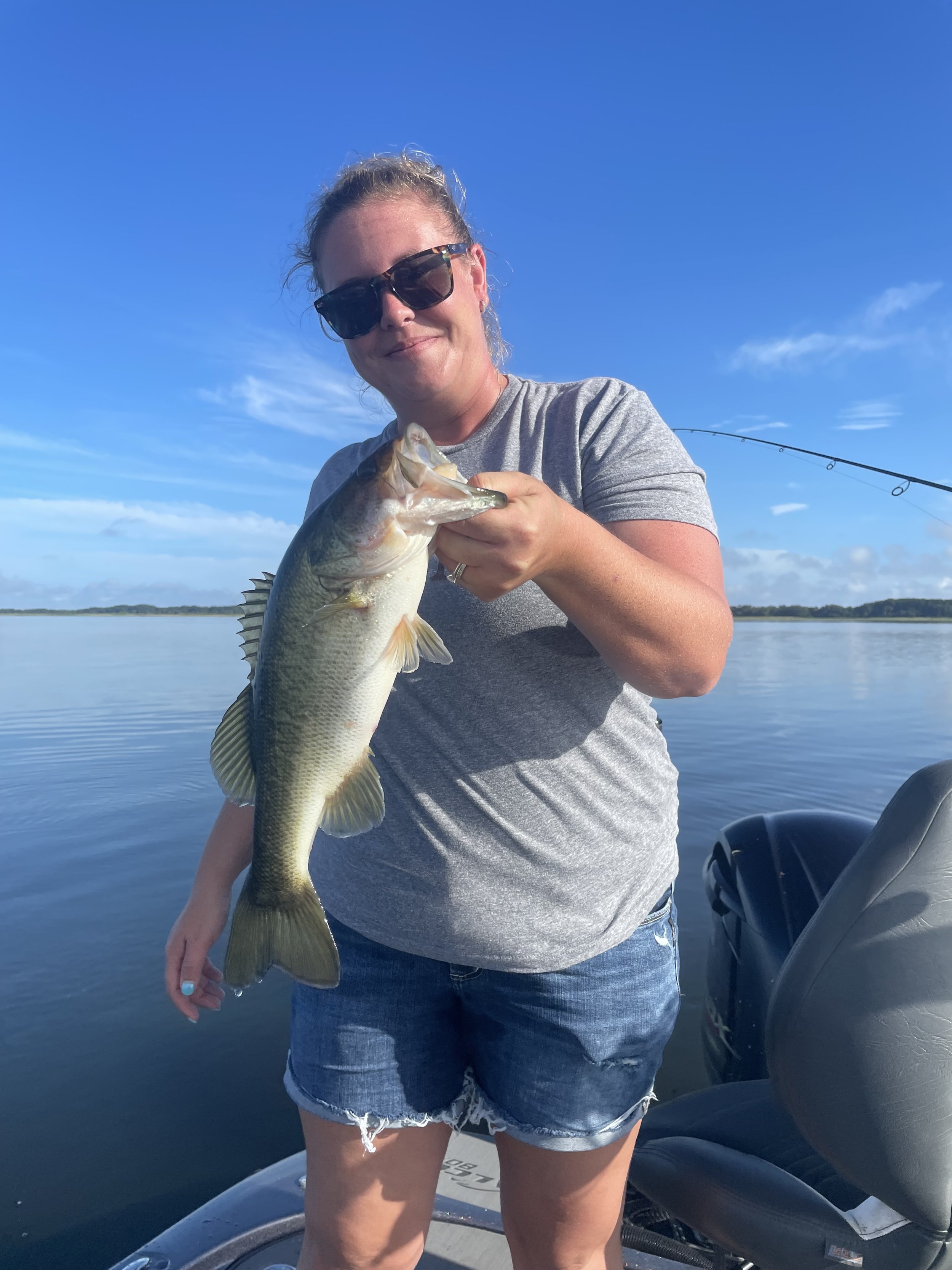 A woman wearing sunglasses, a gray t-shirt, and denim shorts holding a large fish on a boat on a calm lake with a clear blue sky.