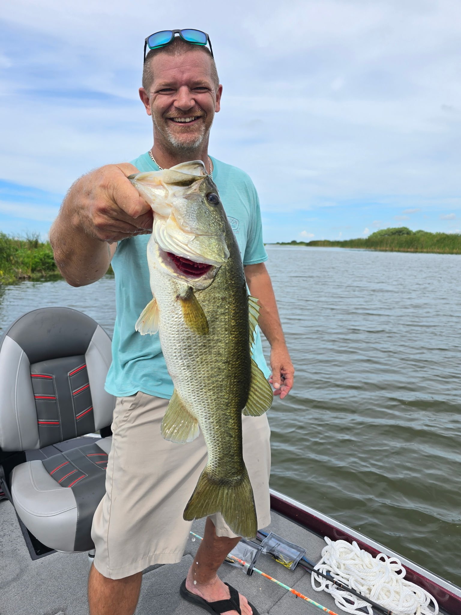 Smiling man on boat holding a large bass fish, with water and greenery in the background.
